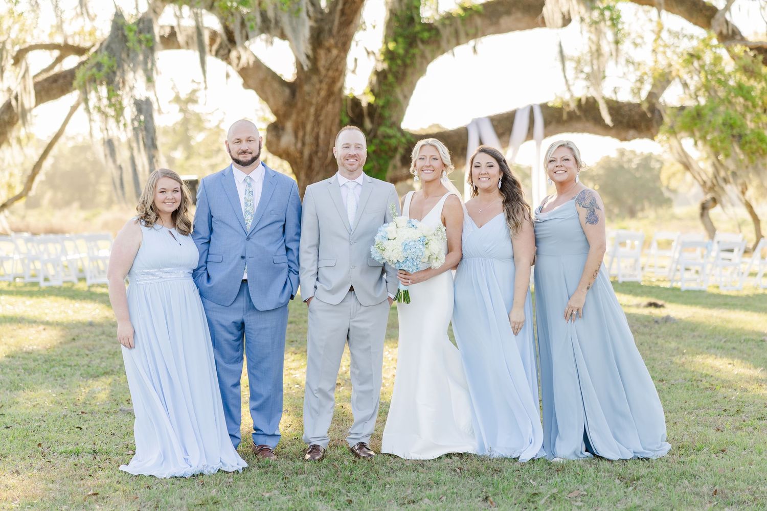 Bride and groom with small wedding party posing under a large oak tree in soft natural light.