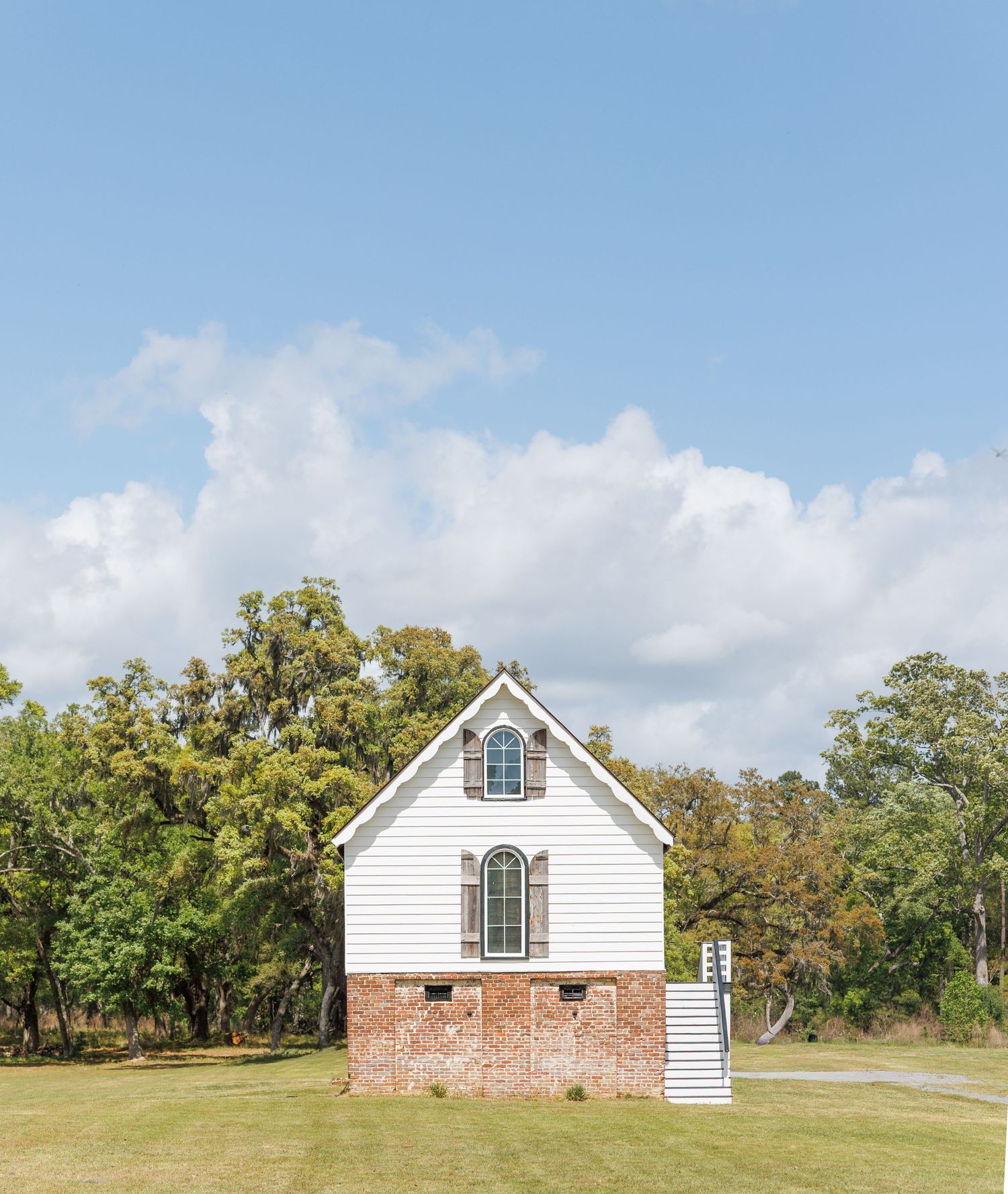 A white historic chapel with brick foundation sits on a green lawn surrounded by trees under a blue sky.