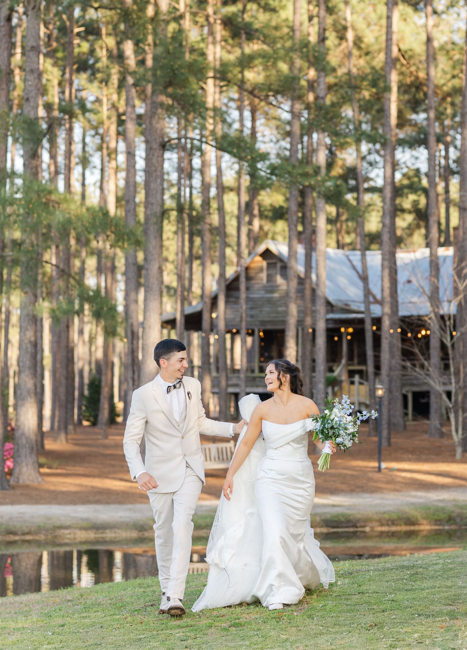 Bride and groom walking hand in hand near pond with rustic wooden barn in pine forest.