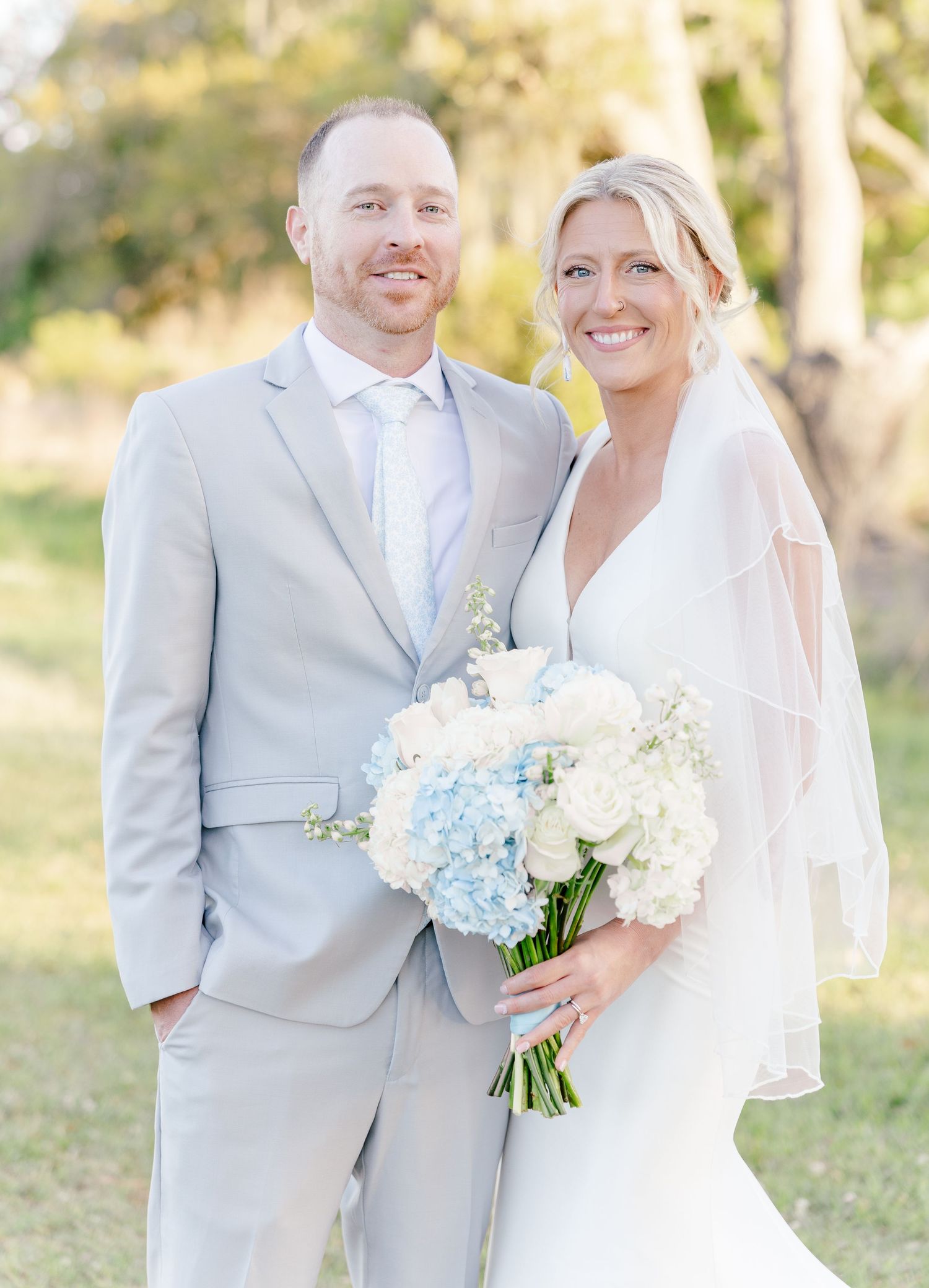 Smiling bride and groom portrait, bride holding white and blue hydrangea bouquet, groom in gray suit.