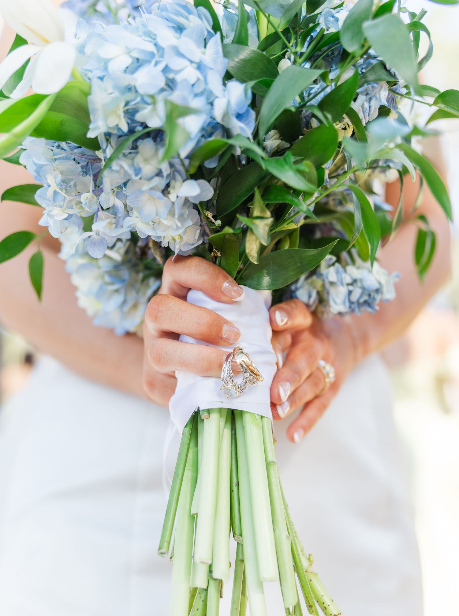 Bride holding blue hydrangea wedding bouquet wrapped in white ribbon with gold diamond engagement ring.