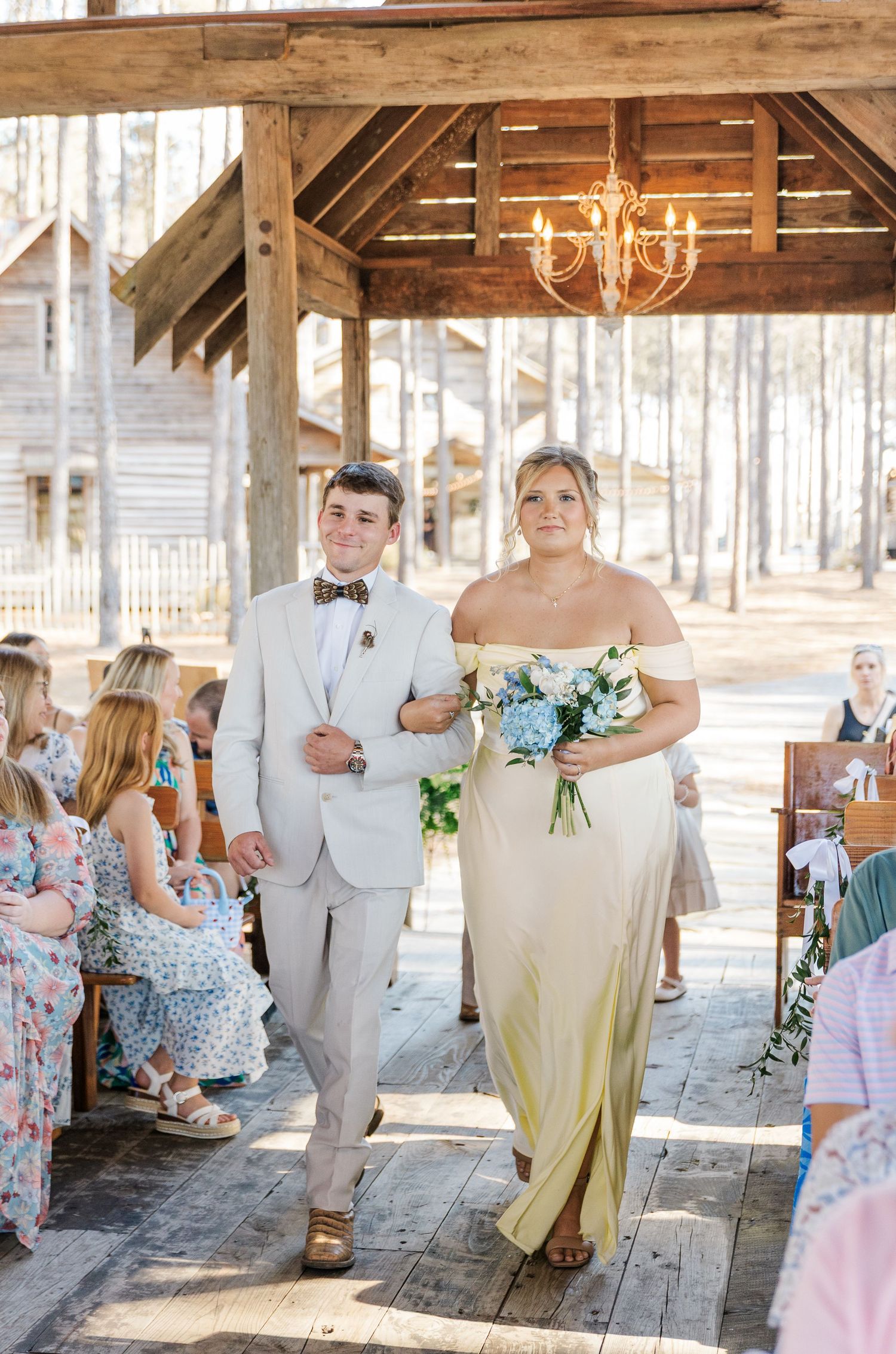 Bridesmaid in yellow gown and groomsman walk down rustic wooden aisle under chandelier at ceremony.
