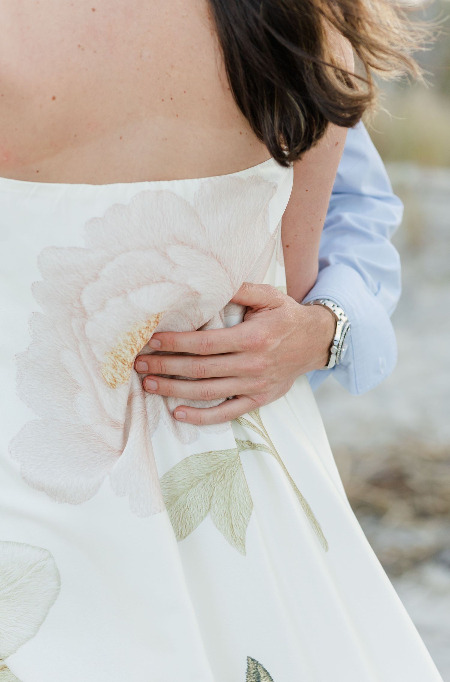 Close-up of a mans hand holding his partners waist in a floral strapless gown during an engagement shoot.