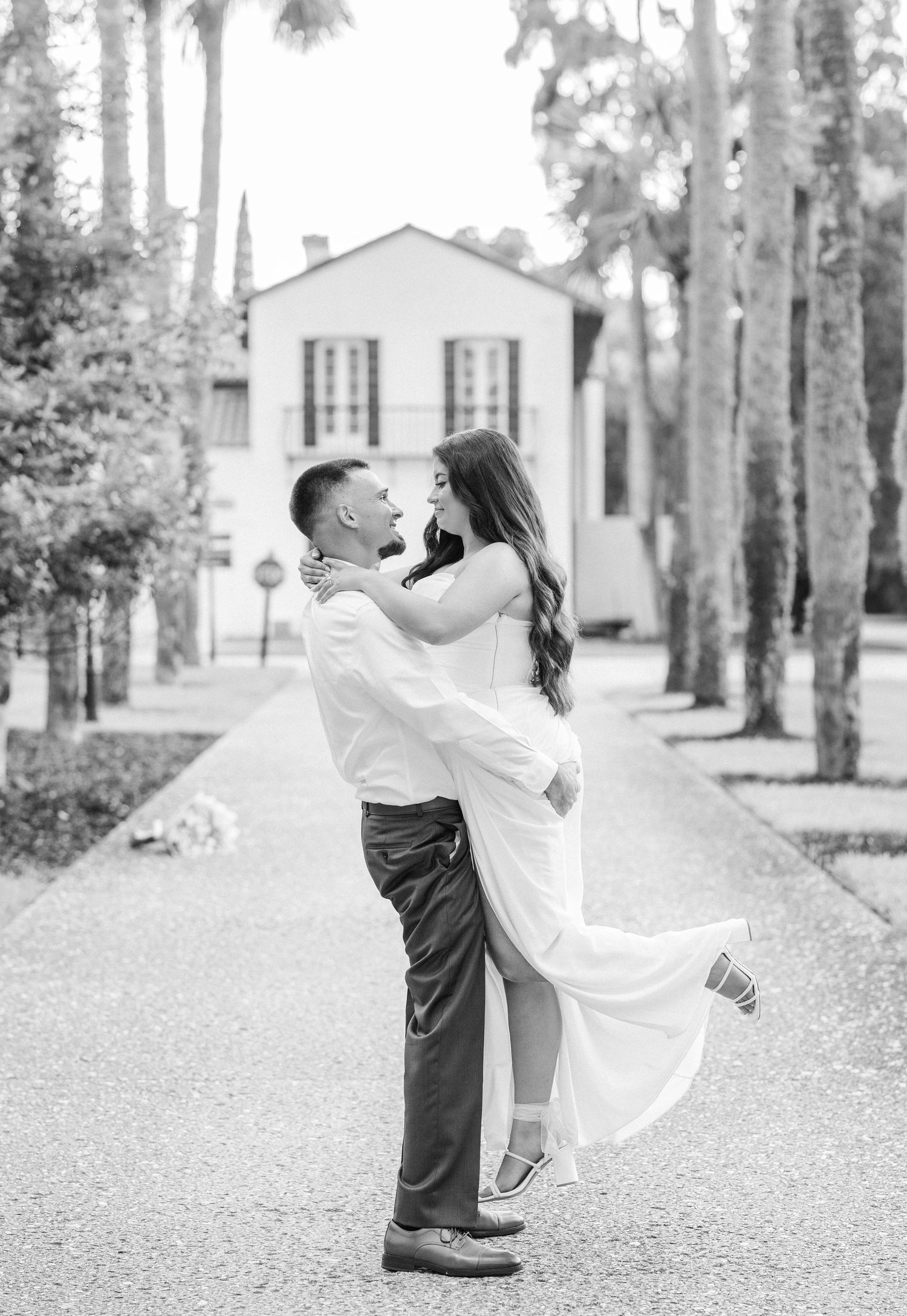 Black and white photo of a groom lifting his bride in a white dress along a palm tree-lined pathway.
