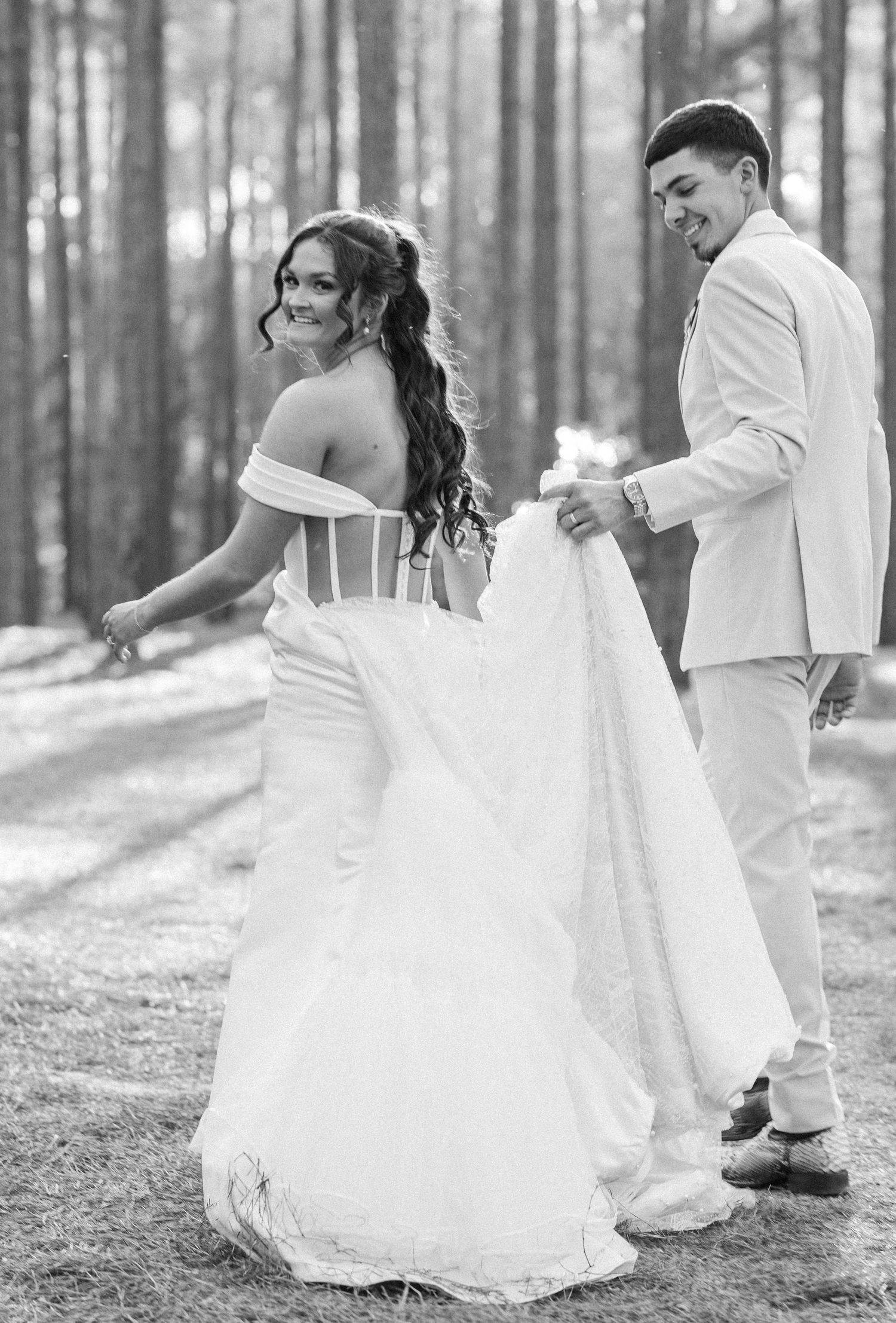 Black and white wedding photo of a couple in white attire standing in a forest setting during their special day.