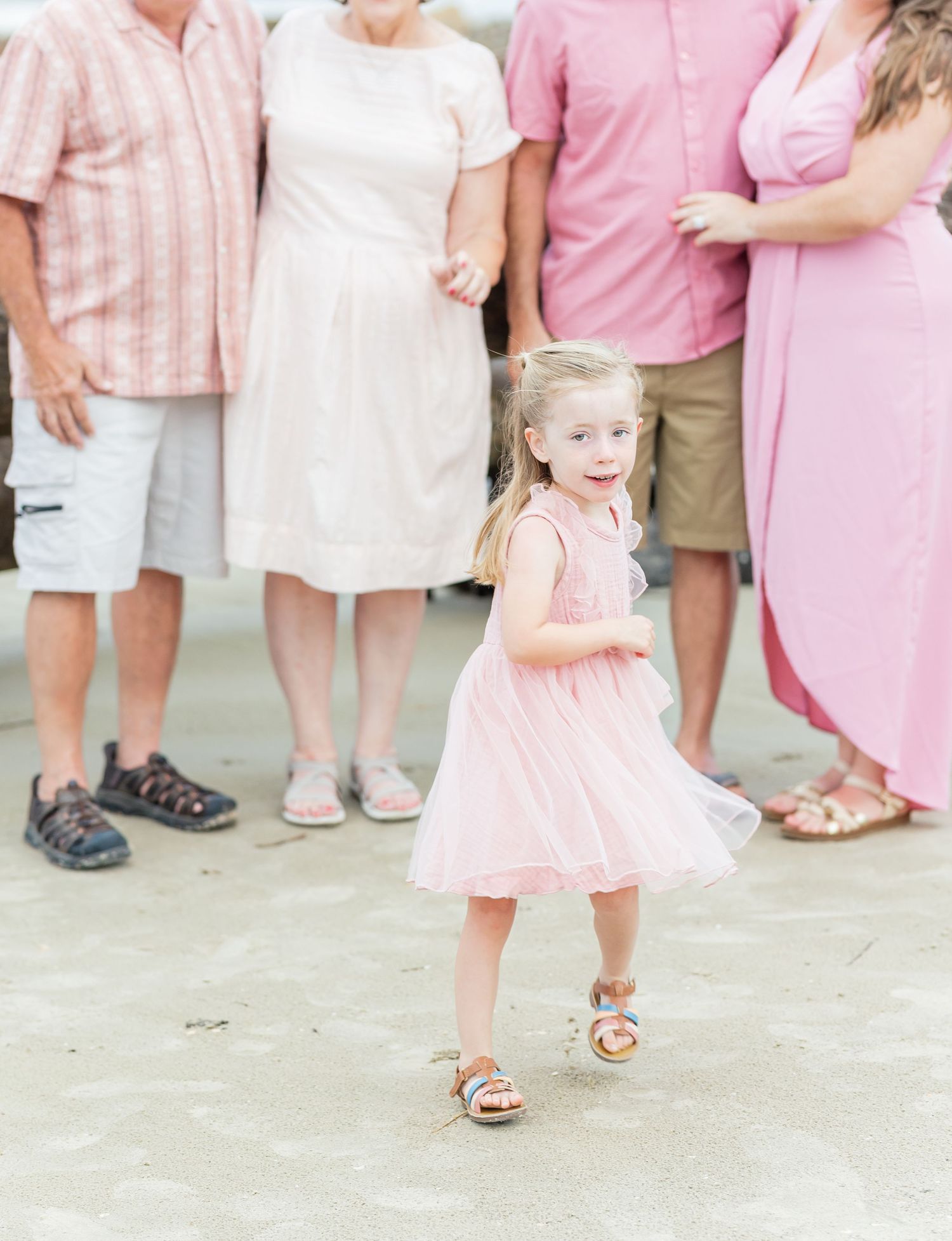 Adorable young girl in a pale pink dress twirls and smiles while standing with others during an outdoor gathering.