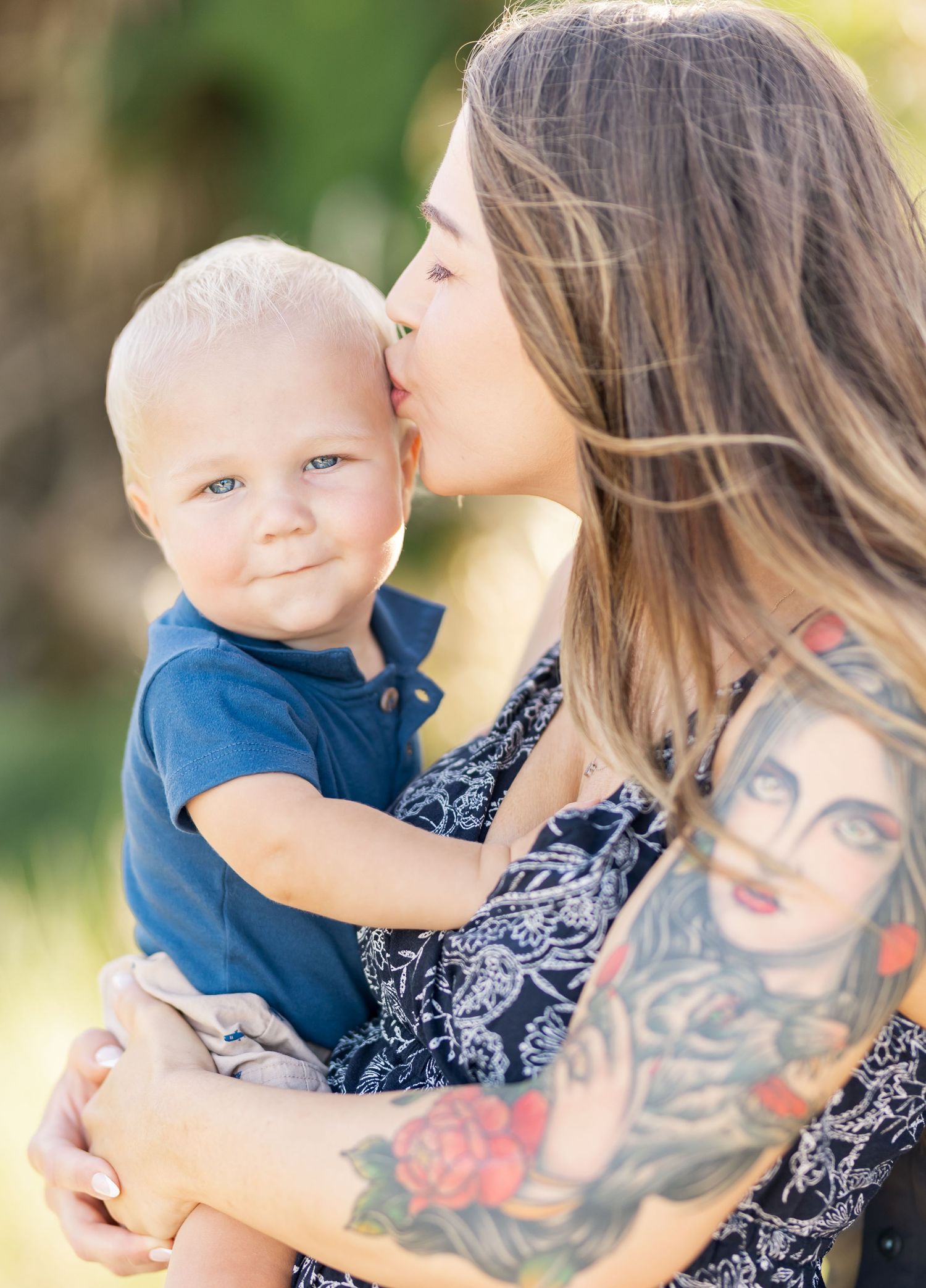 A parent in a navy dress with arm tattoos holds and kisses their small child in a blue outfit against a sunlit background.