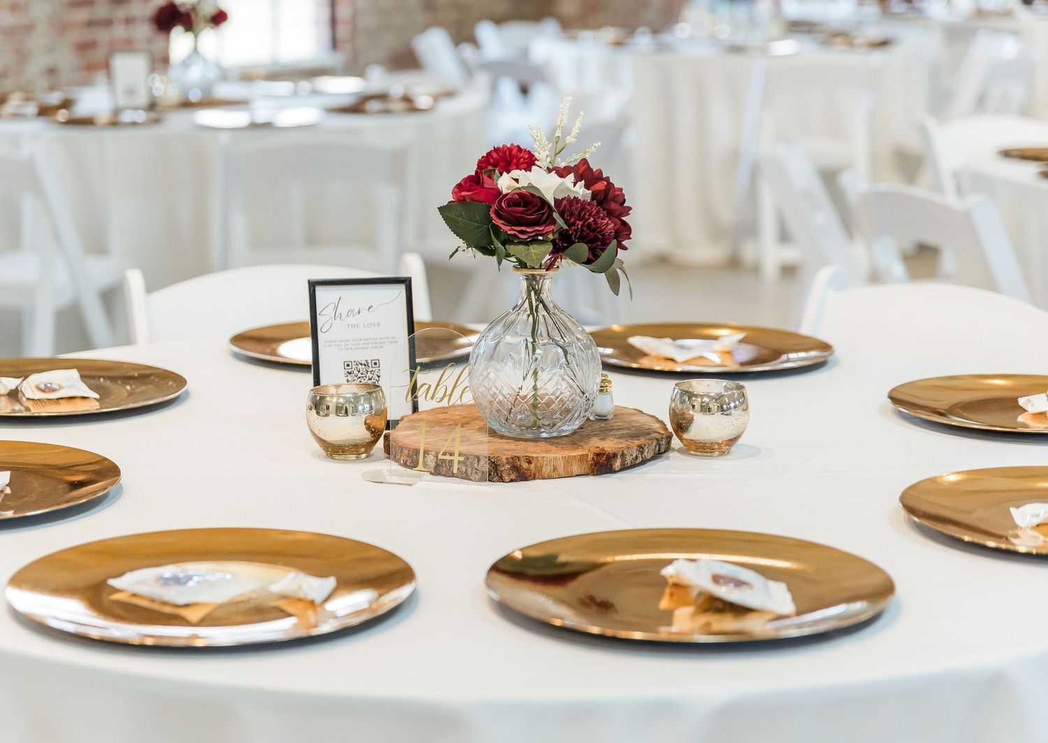 Elegant table setting with gold charger plates and burgundy flower centerpiece on white tablecloth.