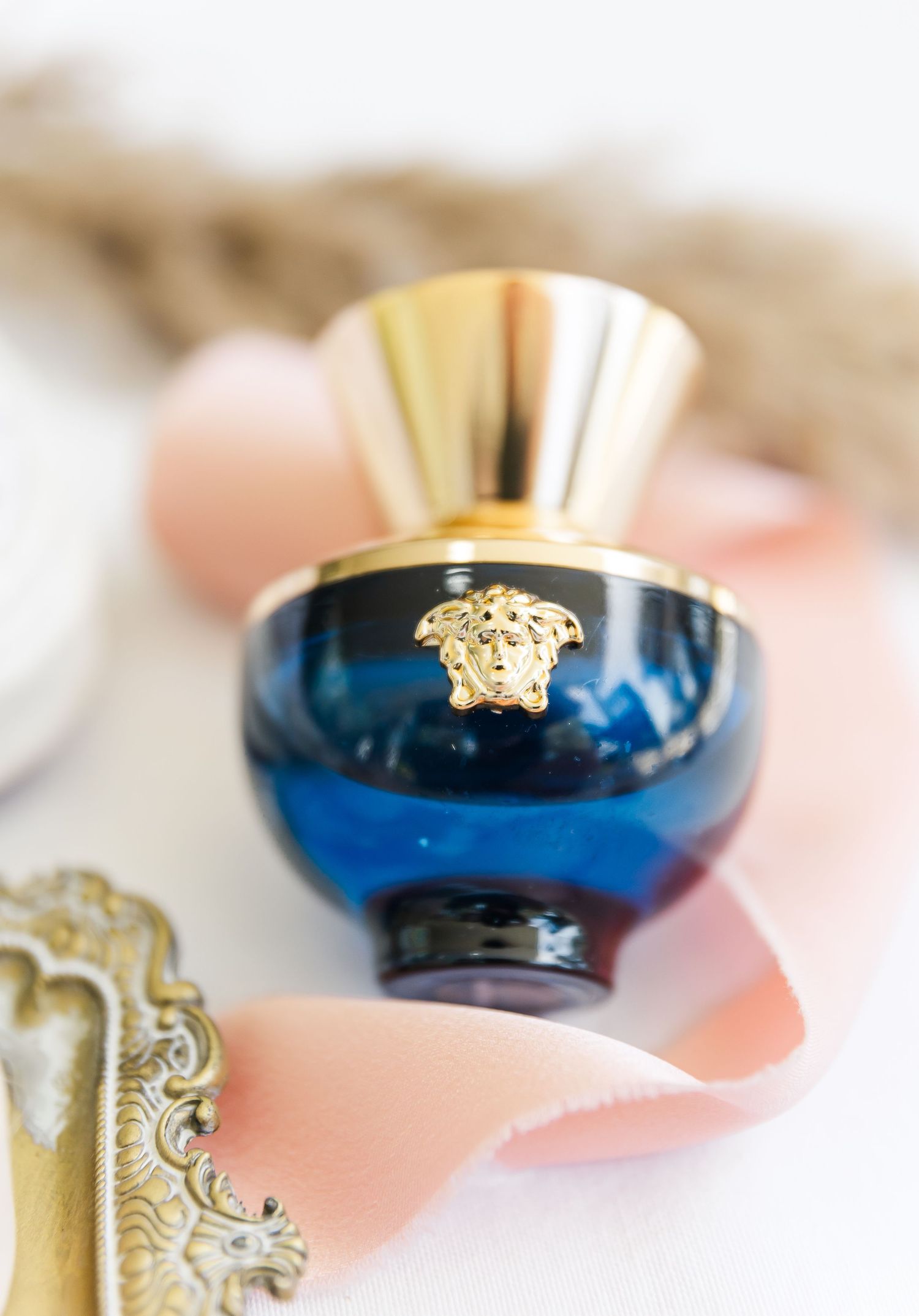 Close-up of an ornate blue and gold jewelry piece with Versace Medusa head logo against a soft pink background.