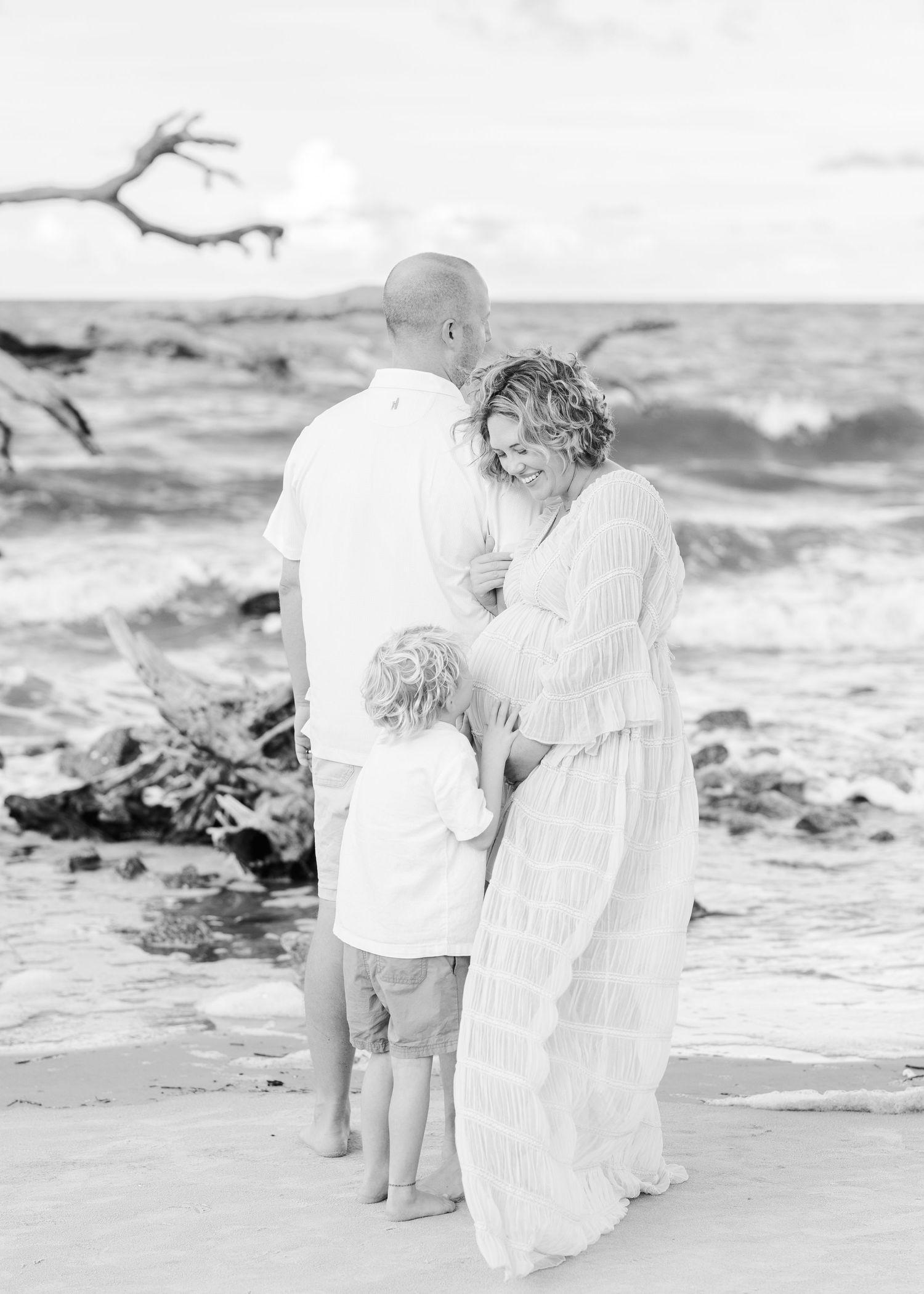 Black and white photo of a family sharing an intimate moment together on a beach with waves crashing behind them.