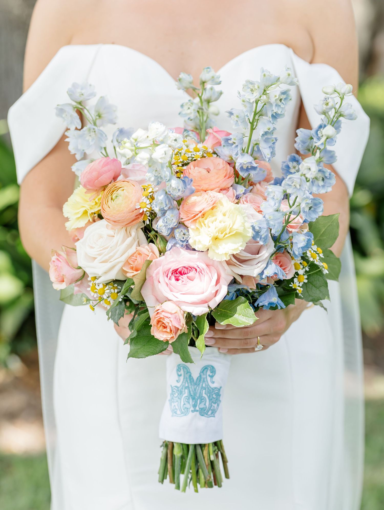 A romantic bridal bouquet featuring pink roses, white roses, blue delphinium, and peach ranunculus with a monogrammed ribbon.