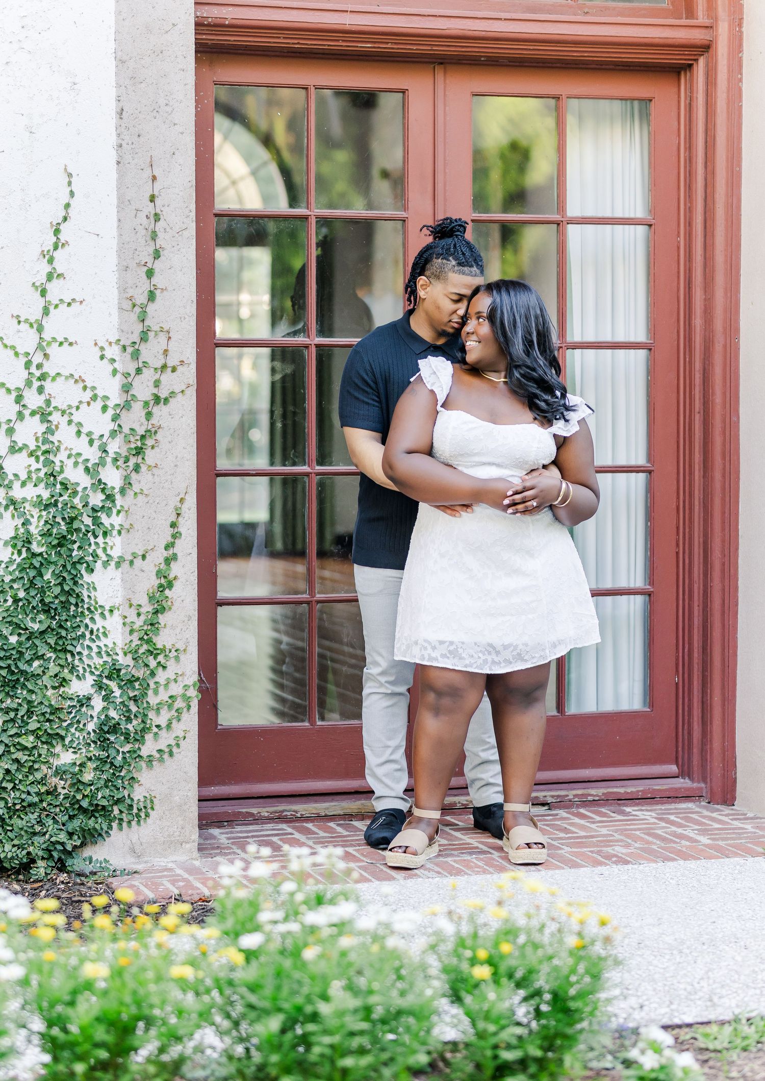 Couple shares intimate moment by red doorway surrounded by greenery.