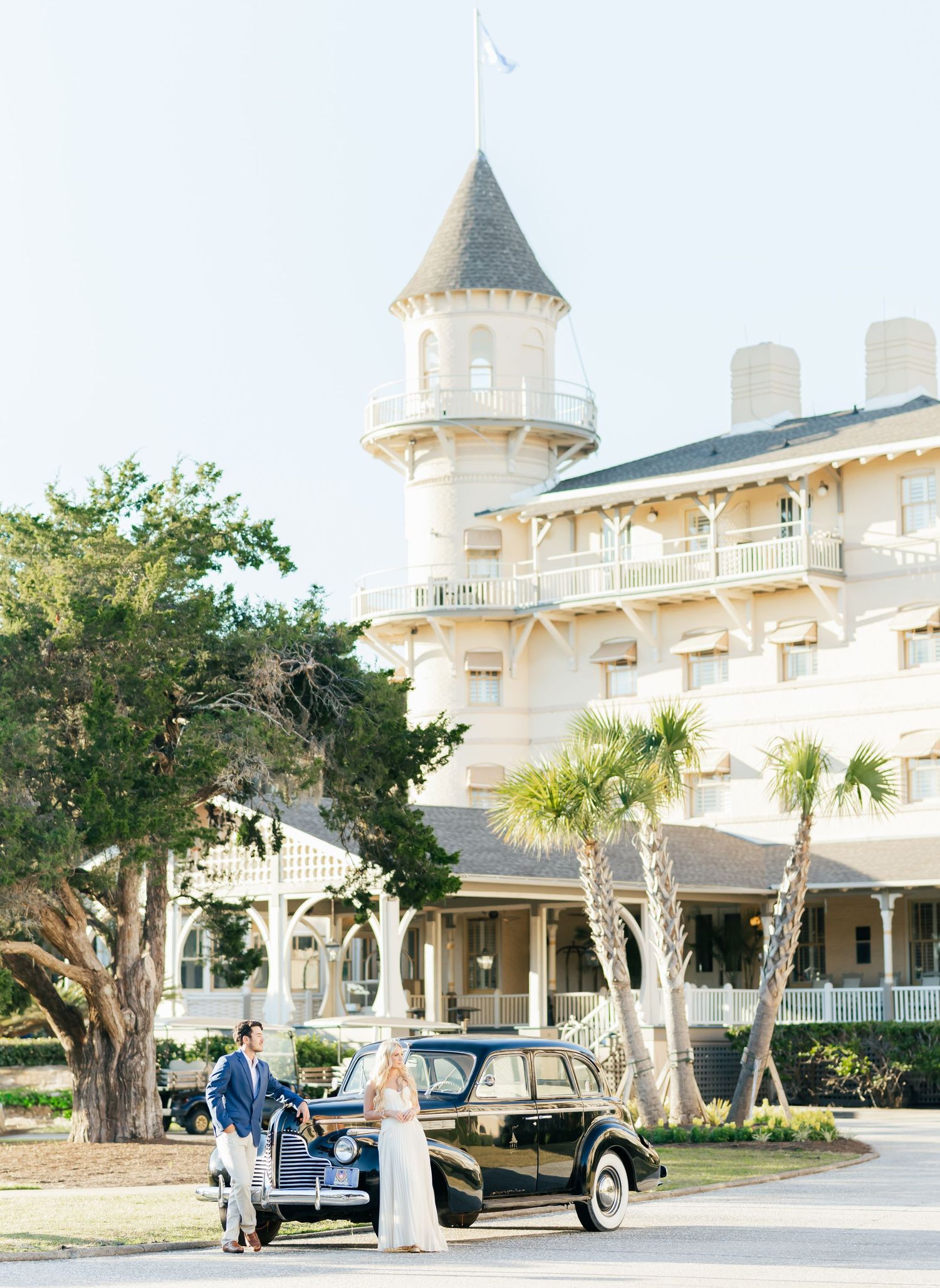 Elegant Victorian-style mansion with white exterior and turrets photographed with a vintage car in front during sunset.