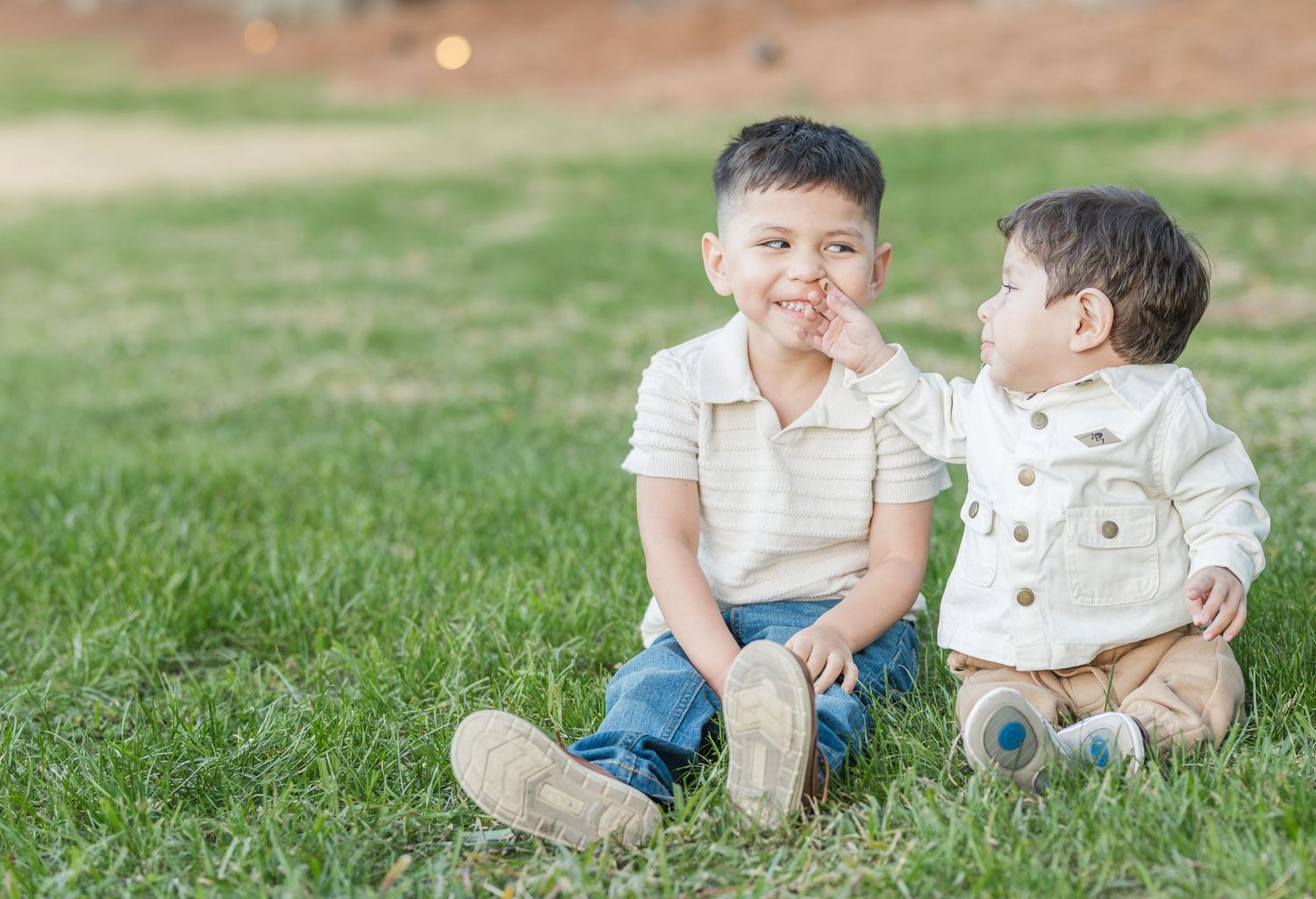 Two children sit together on green grass while playing and interacting during an outdoor photo session.