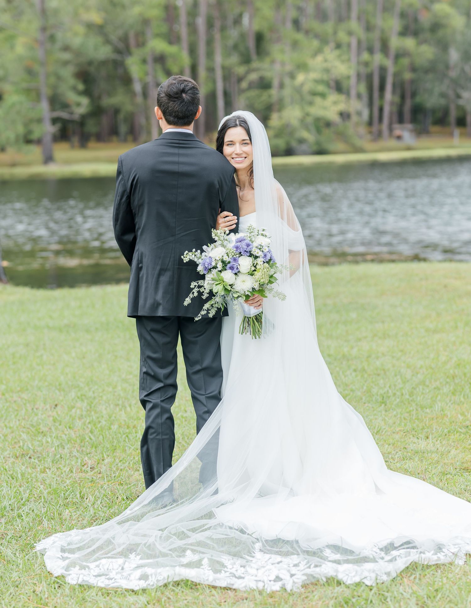 Scenic lakeside wedding photography session with couple in white dress and black suit on green lawn.