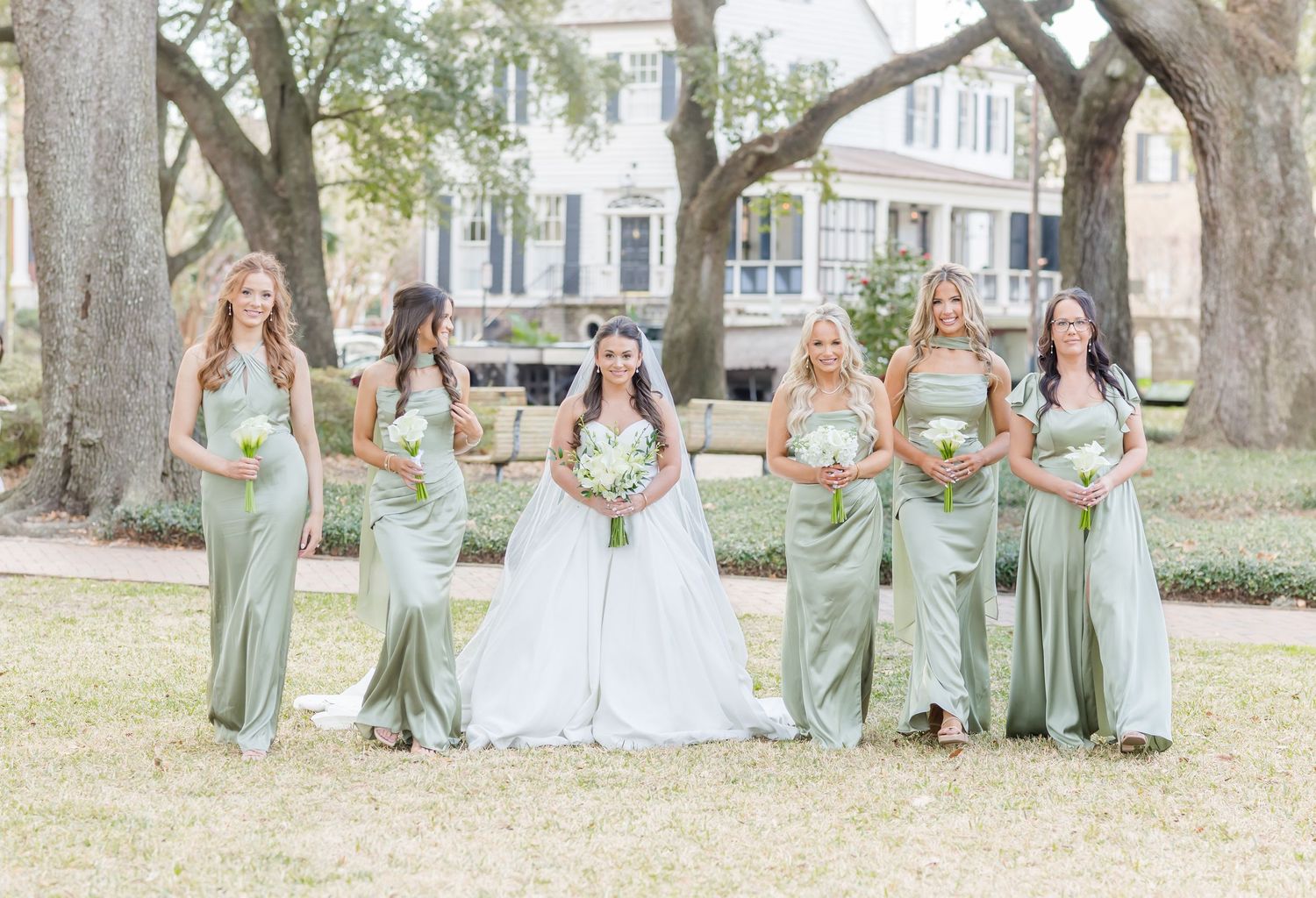 Bridesmaids in sage green dresses pose together outdoors on a sunny day at a wedding.