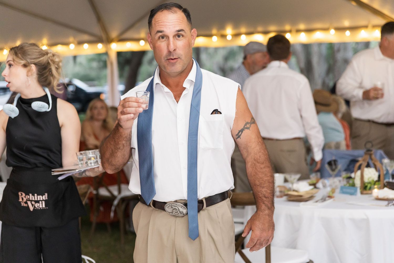 Wedding guests dance and celebrate at an outdoor reception under a white tent with string lights.