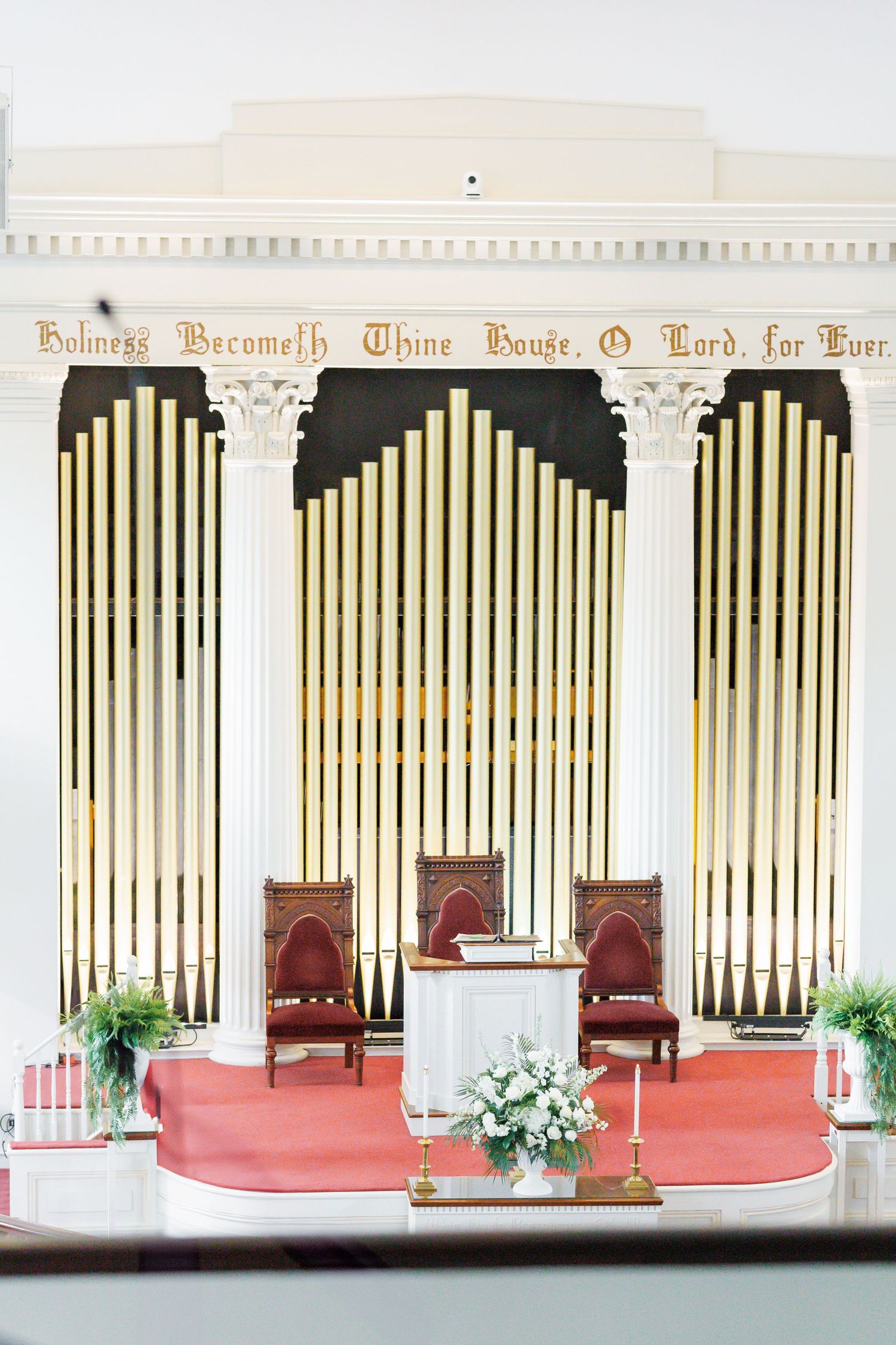 Church altar with white organ pipes, red chairs, and floral arrangements in an elegant sanctuary setting.