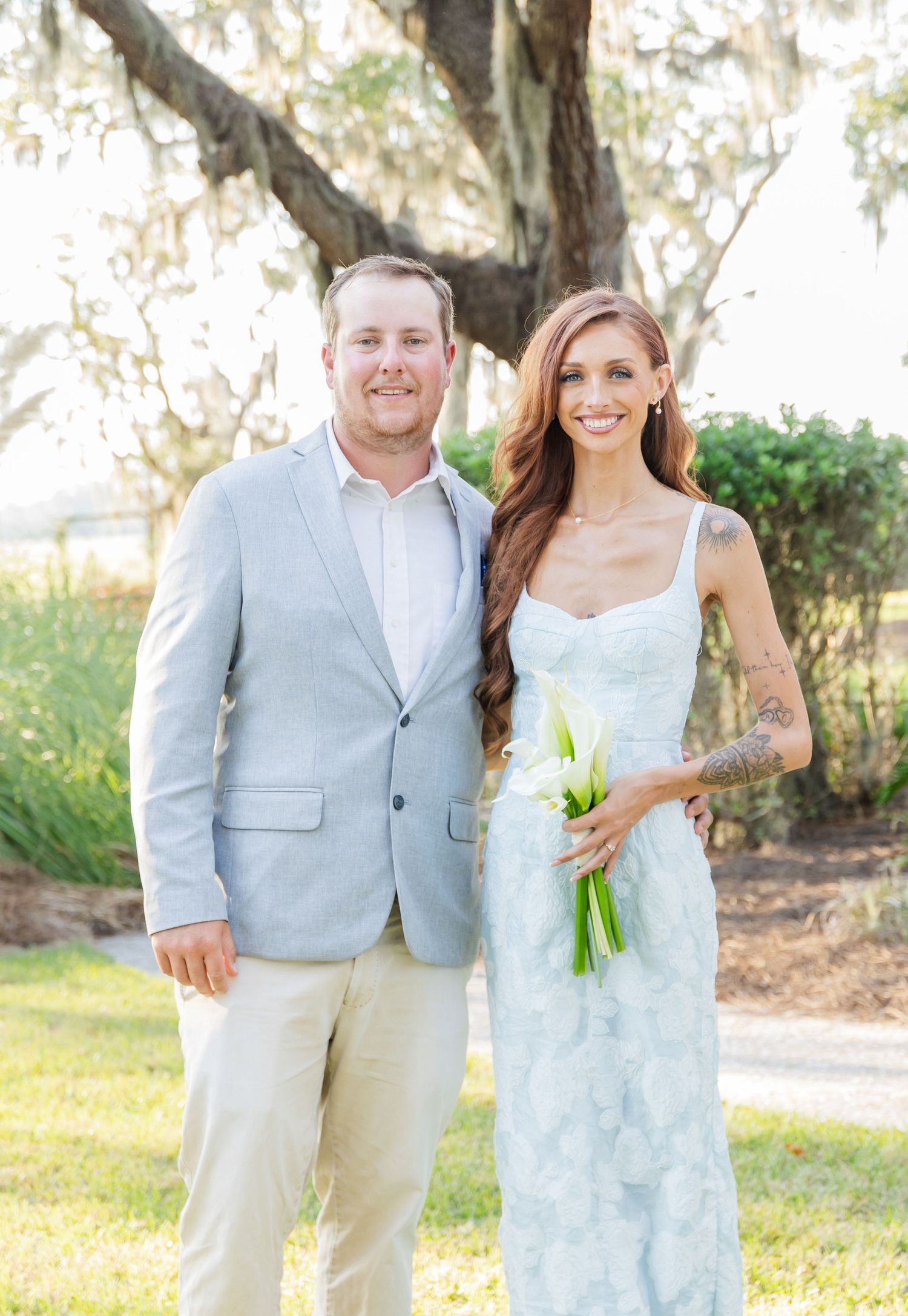 A couple in light blue and white attire poses together outdoors under Spanish moss-draped trees.