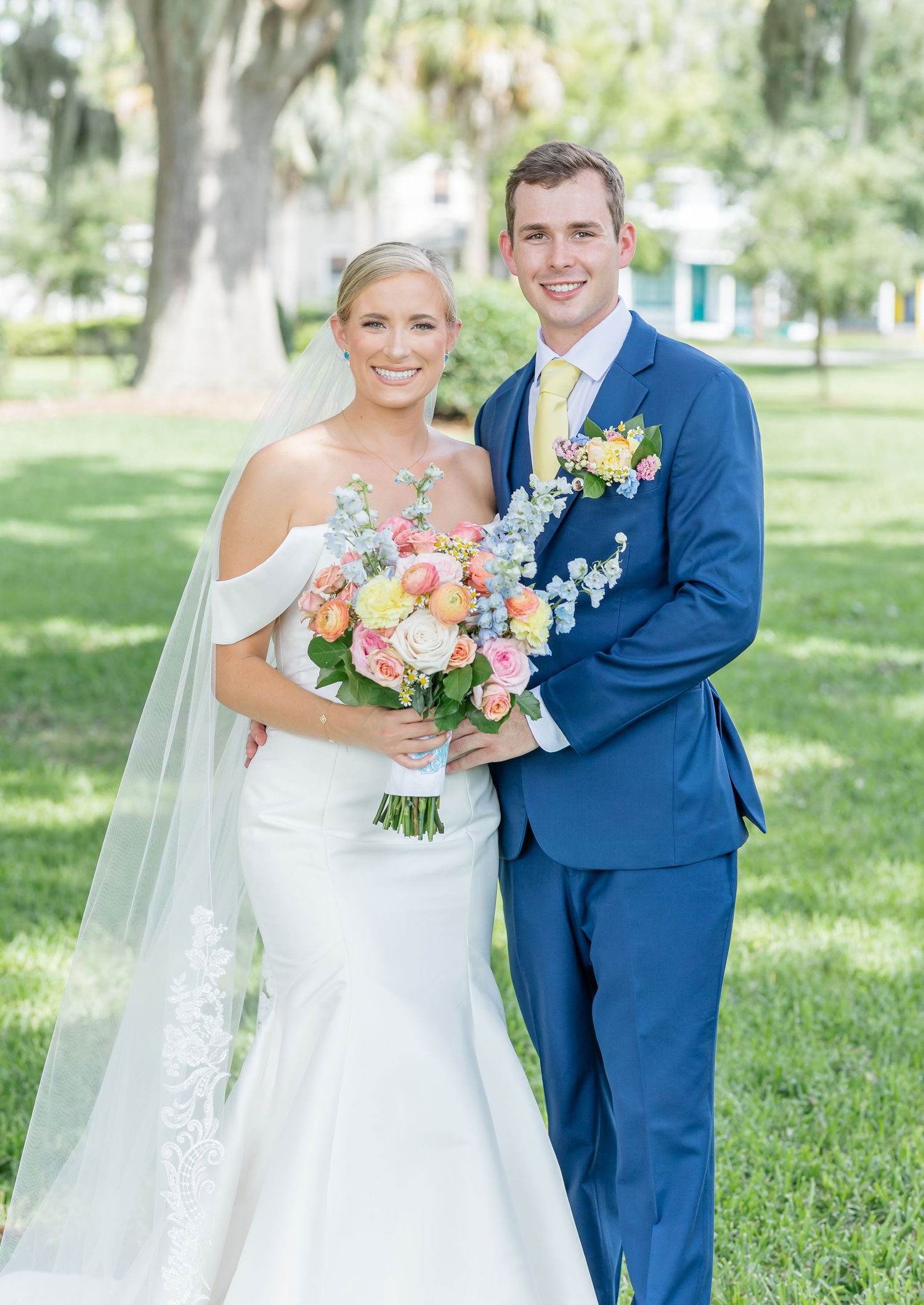 A couple in wedding attire poses outdoors, with the bride in white and groom in navy blue holding a colorful bouquet.