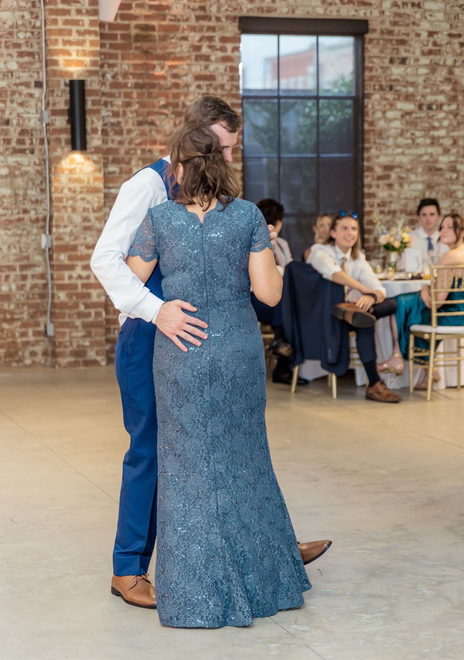 A couple dances together in a rustic brick venue, with the person wearing a blue lace dress and their partner in navy.