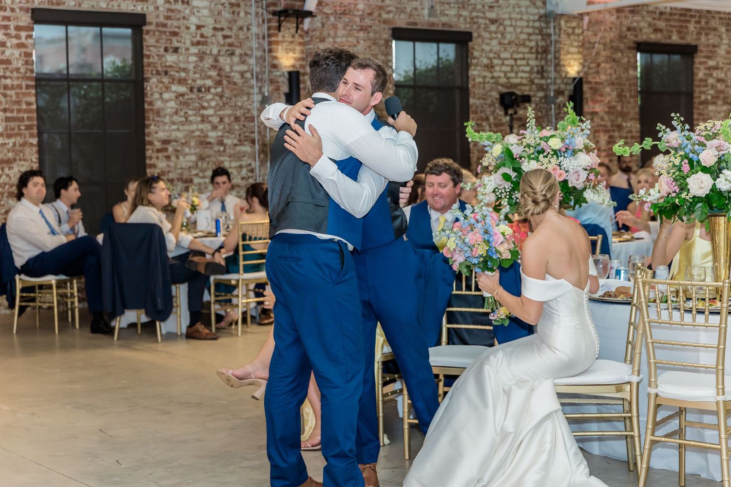 A couple dances together at a wedding reception in a rustic brick venue with floral arrangements and gold chairs.