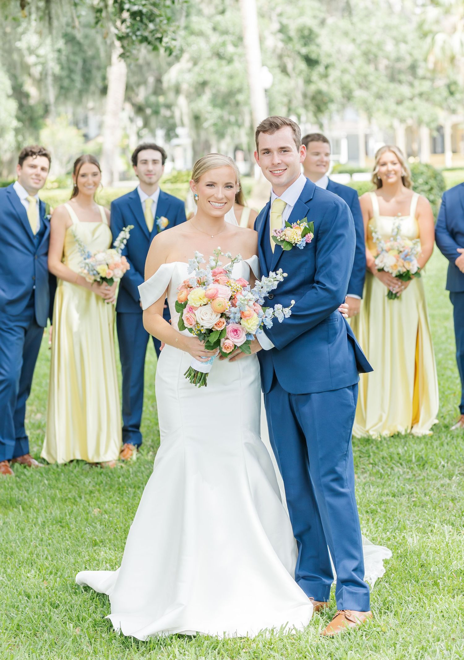Wedding party poses outdoors with couple in center wearing navy suits and yellow dresses under Spanish moss trees.