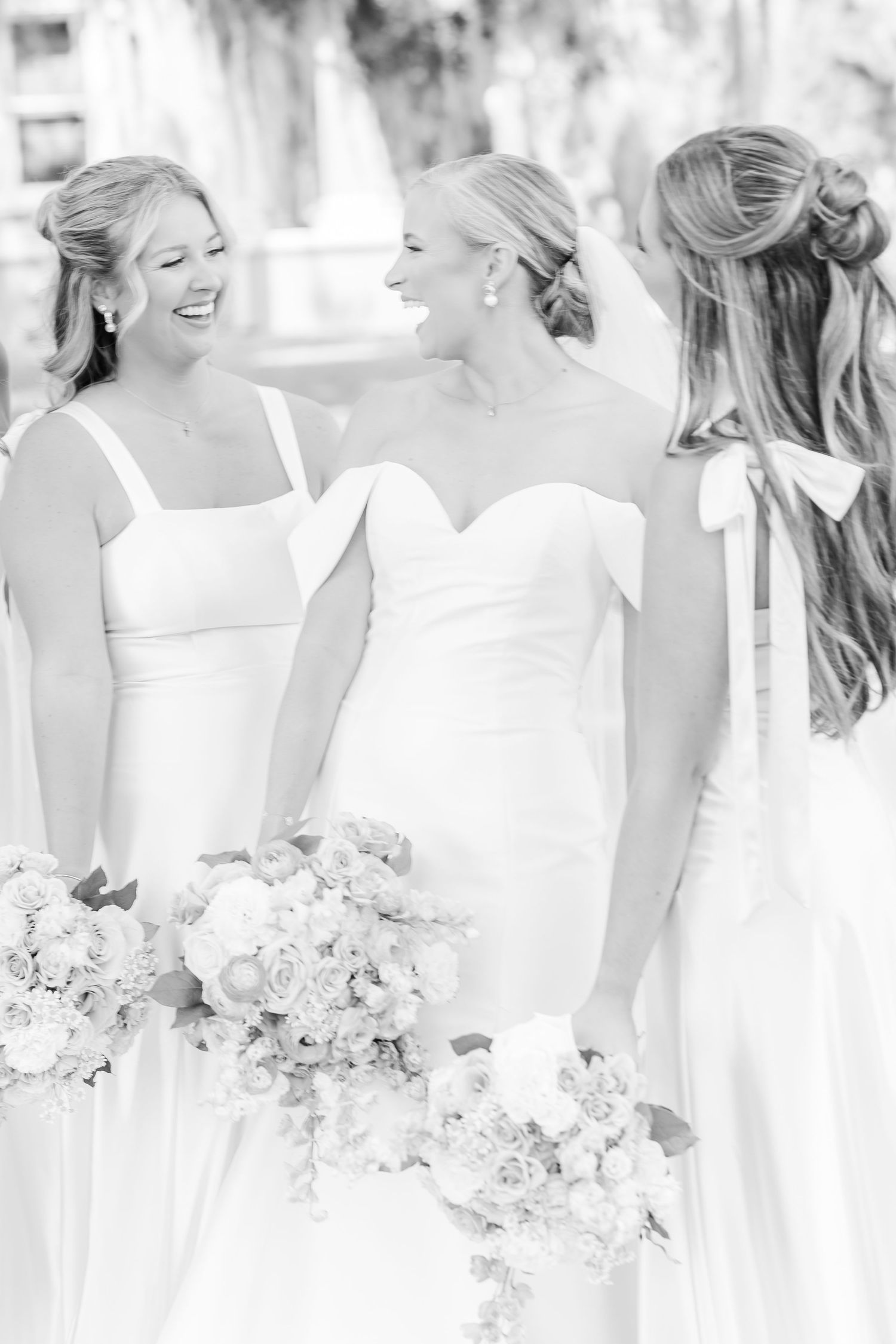 Row of bridesmaids in elegant white dresses holding bouquets against a light background.
