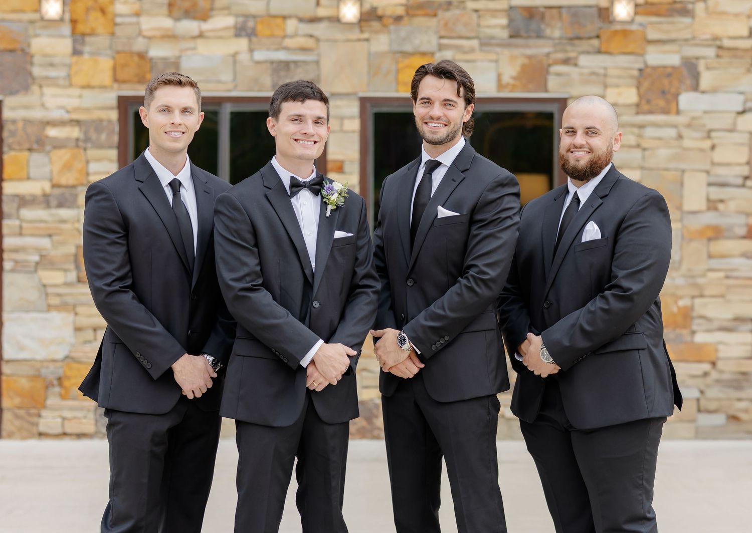 Four groomsmen in formal black tuxedos and suits pose together against a stone wall backdrop.