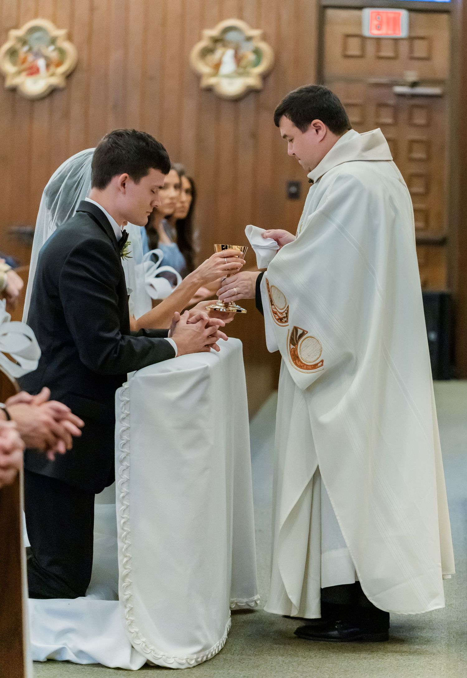 Clergy members in white robes and people in dark suits gather in a church setting for a ceremony.