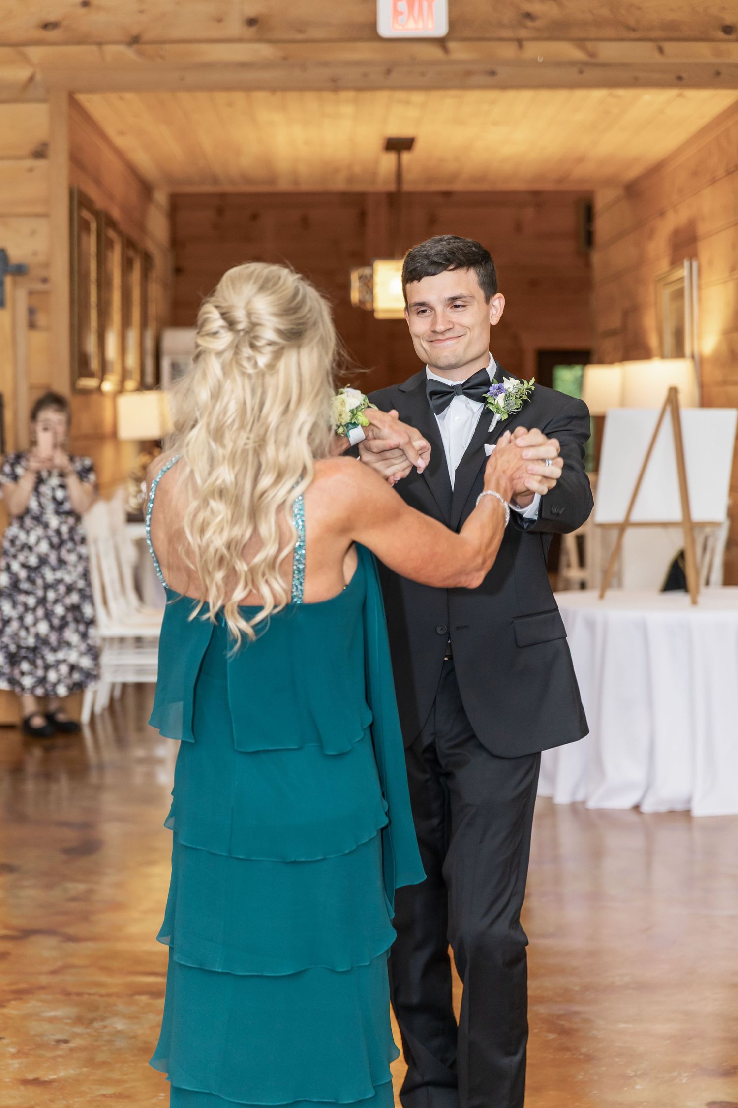 A couple in formal attire share a dance in a rustic wooden venue with white decor.