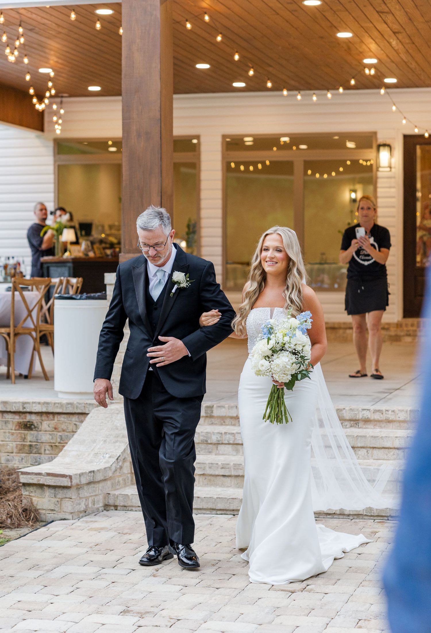 Smiling bride in white strapless gown being escorted down the aisle by her father at outdoor wedding ceremony.