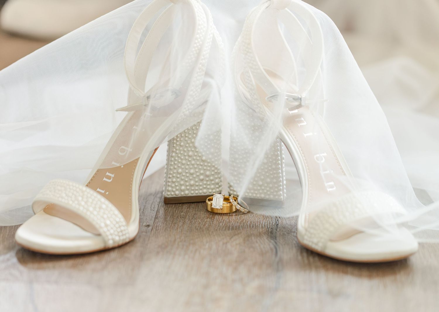 Elegant pearl-embellished white bridal heels with ankle straps beside gold wedding rings on a wooden floor.