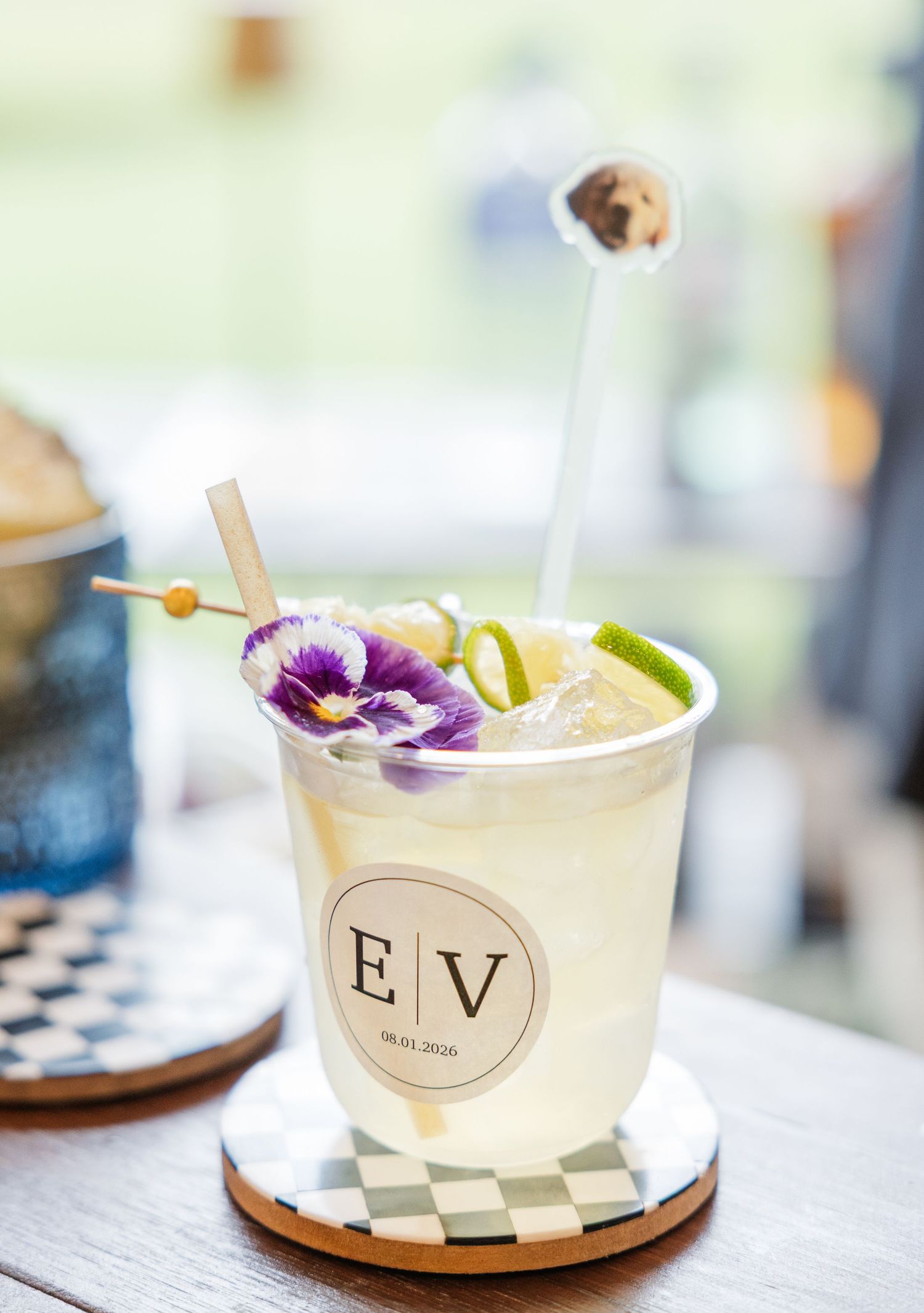 Elegant wedding cocktail in a custom monogrammed cup with purple flower garnish and lime on a checkered coaster.