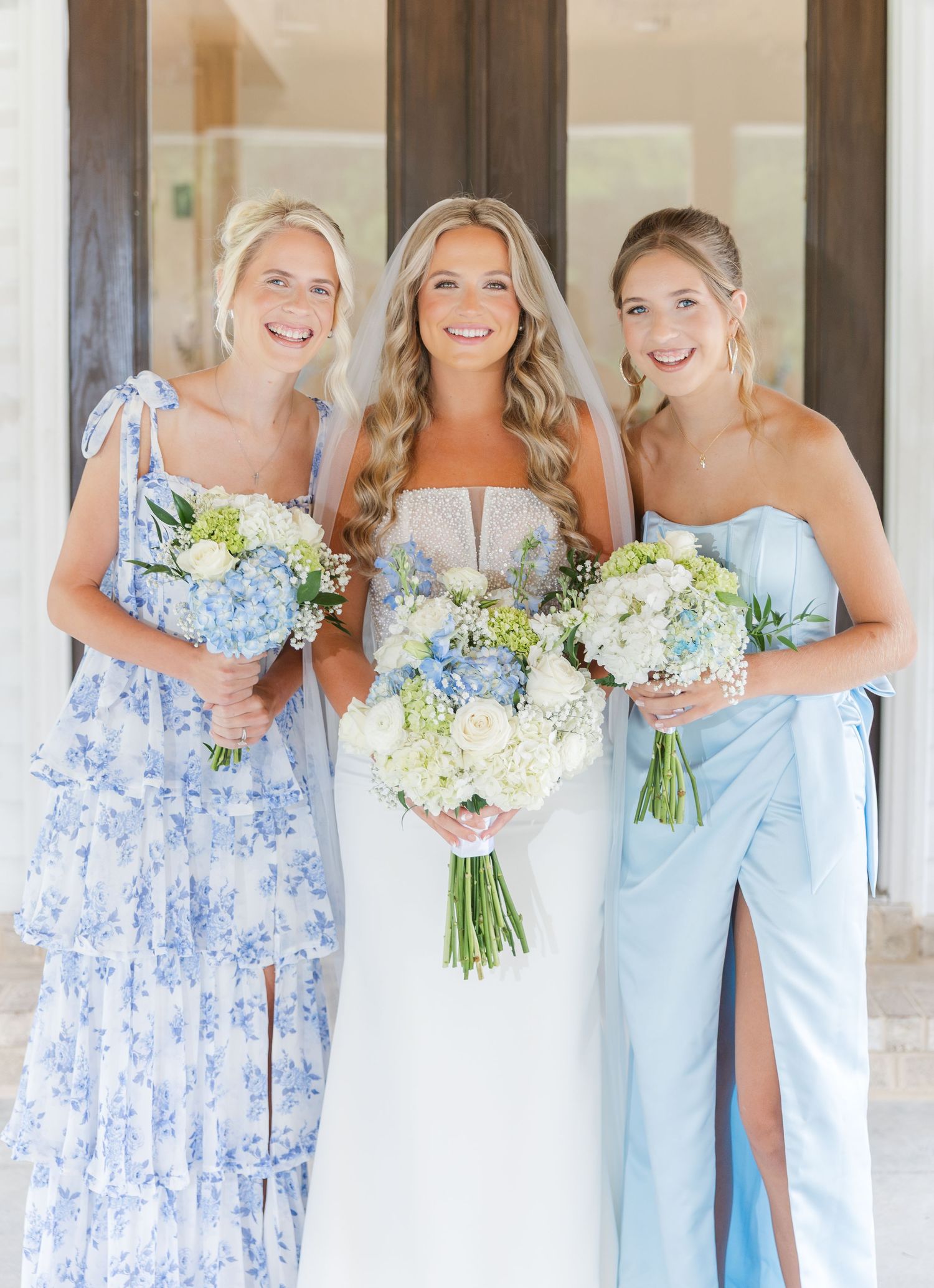 Bride in white gown with two bridesmaids in blue dresses holding white and blue floral bouquets outdoors.