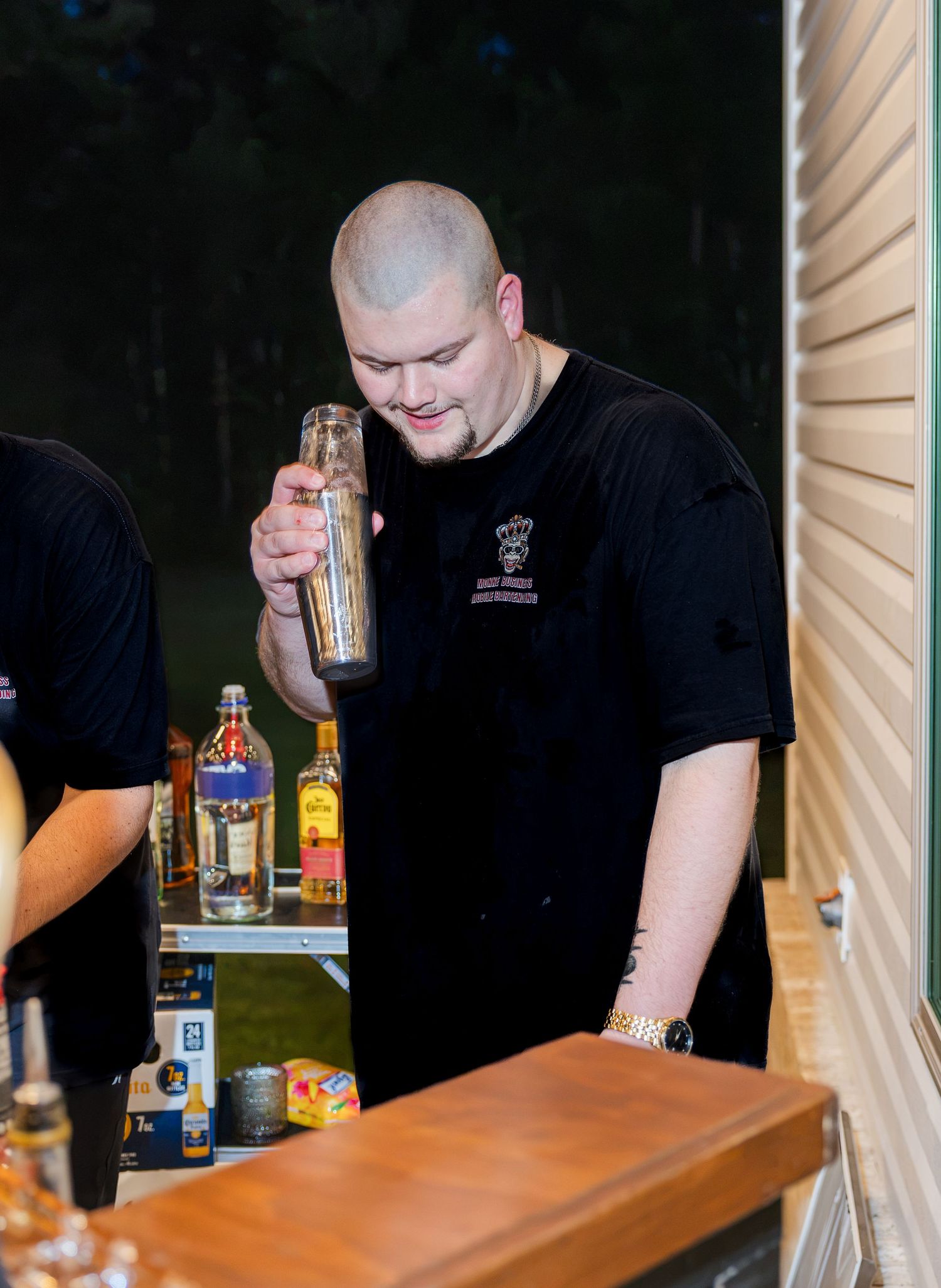 Male bartender in black uniform shaking cocktail shaker at outdoor wedding bar at night.