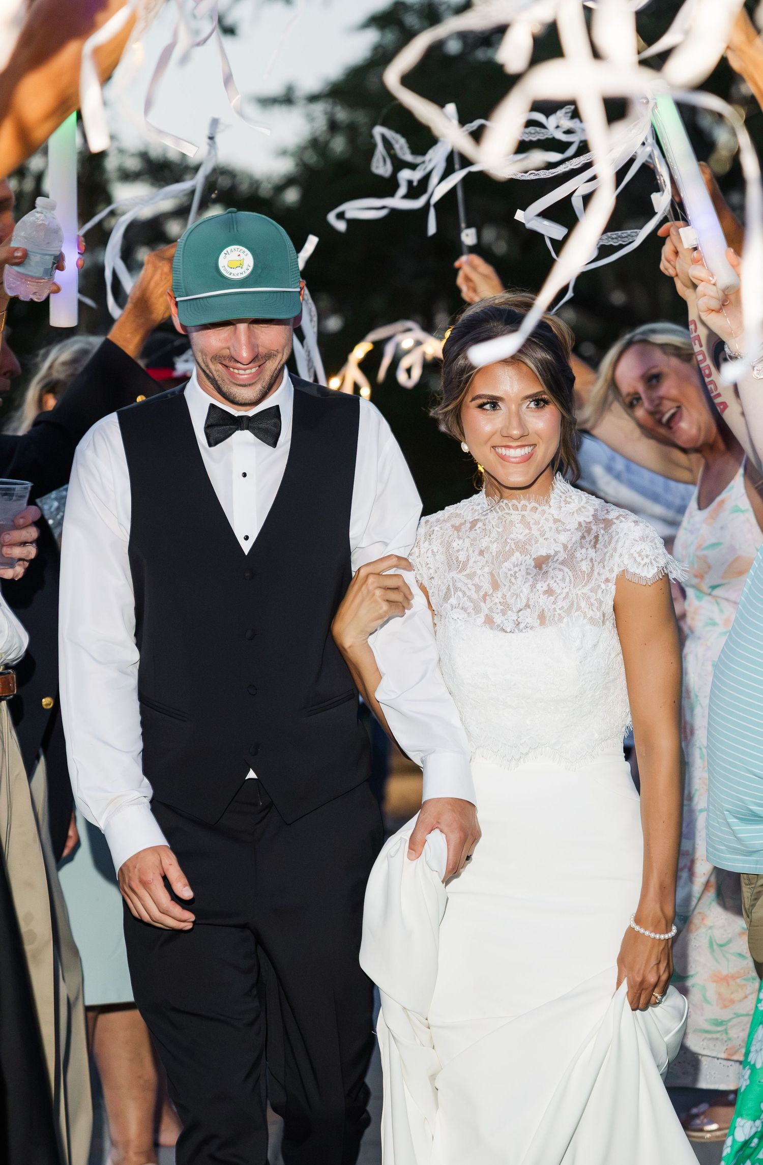 Bride in lace gown and groom in tuxedo with green cap walk through ribbon send-off at outdoor wedding reception.