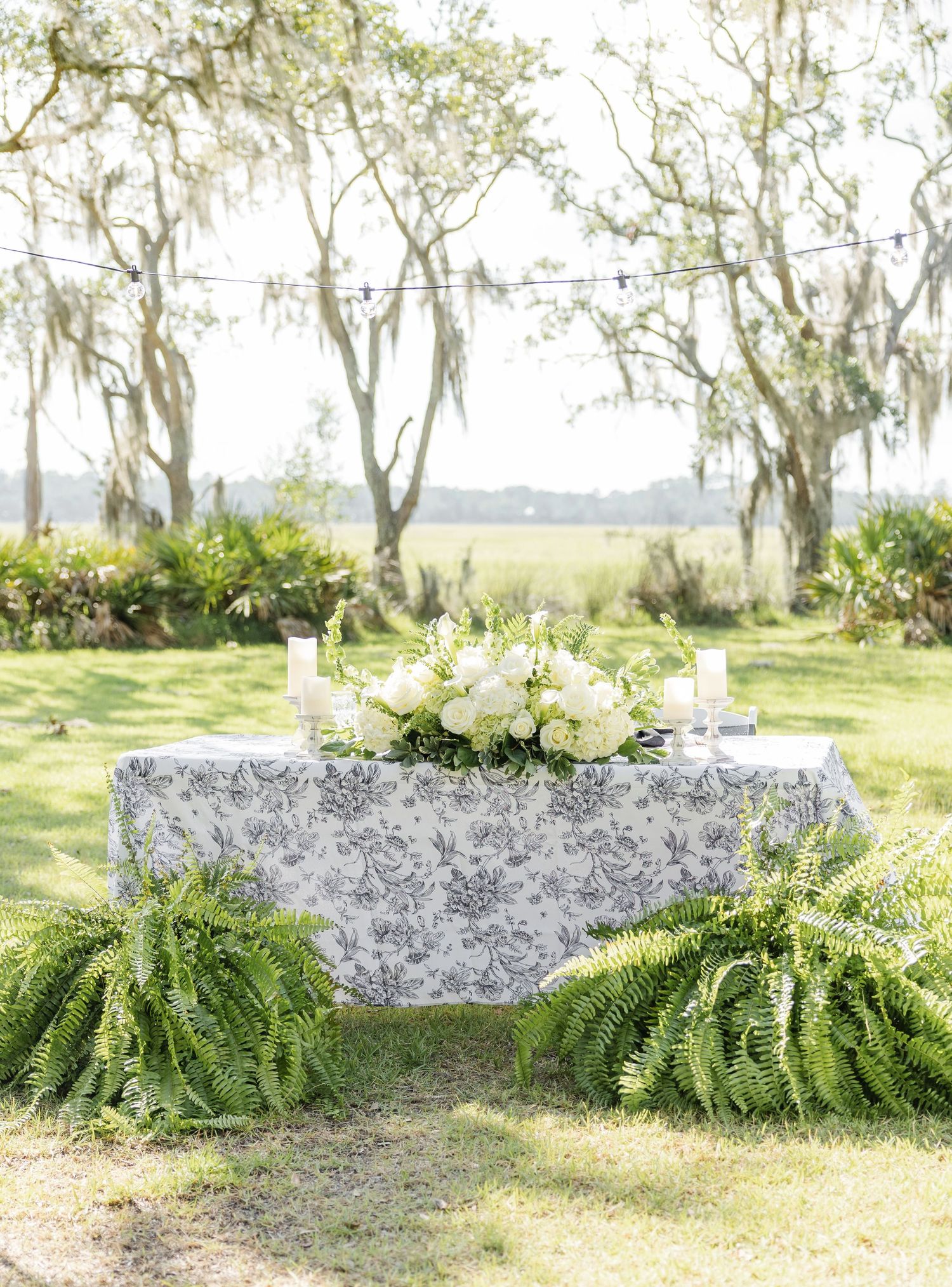 Outdoor wedding sweetheart table with floral toile linen, white roses, candles, and fern garland beneath moss-draped trees.