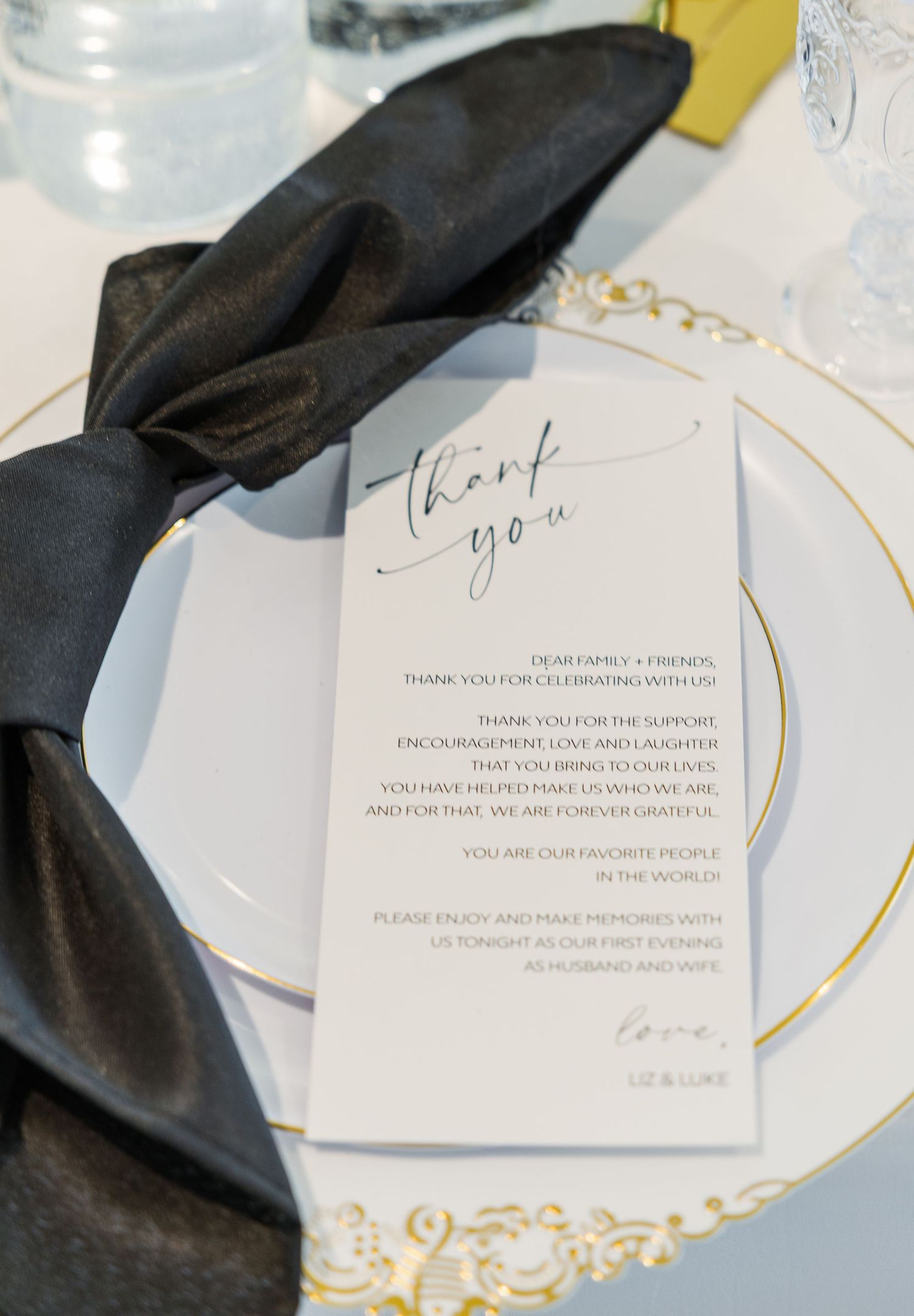 Elegant wedding thank you card on gold-rimmed charger plate with black satin napkin, signed by Liz and Luke.