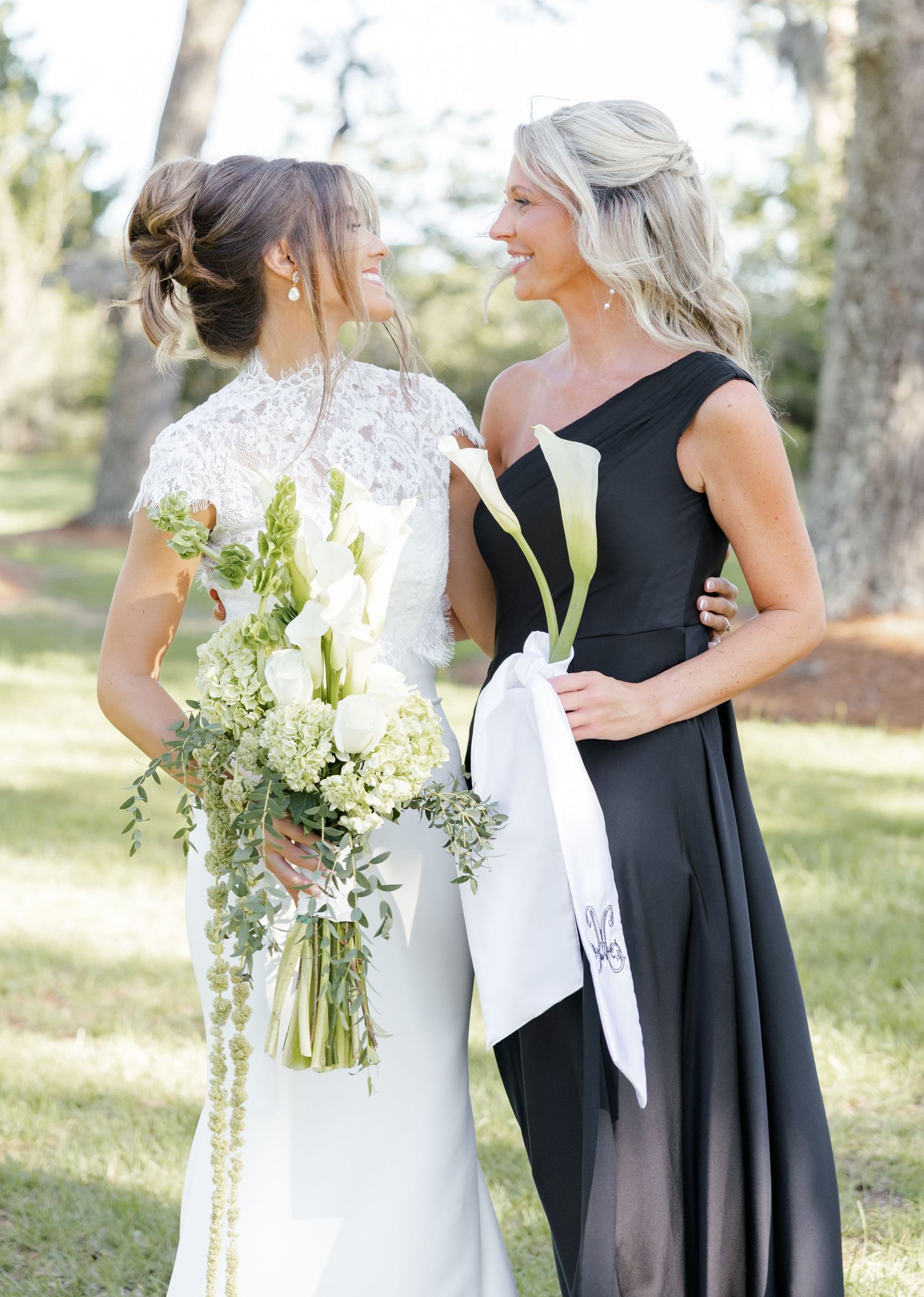 Bride in lace gown holding white floral bouquet stands with bridesmaid in black one-shoulder dress holding calla lilies.