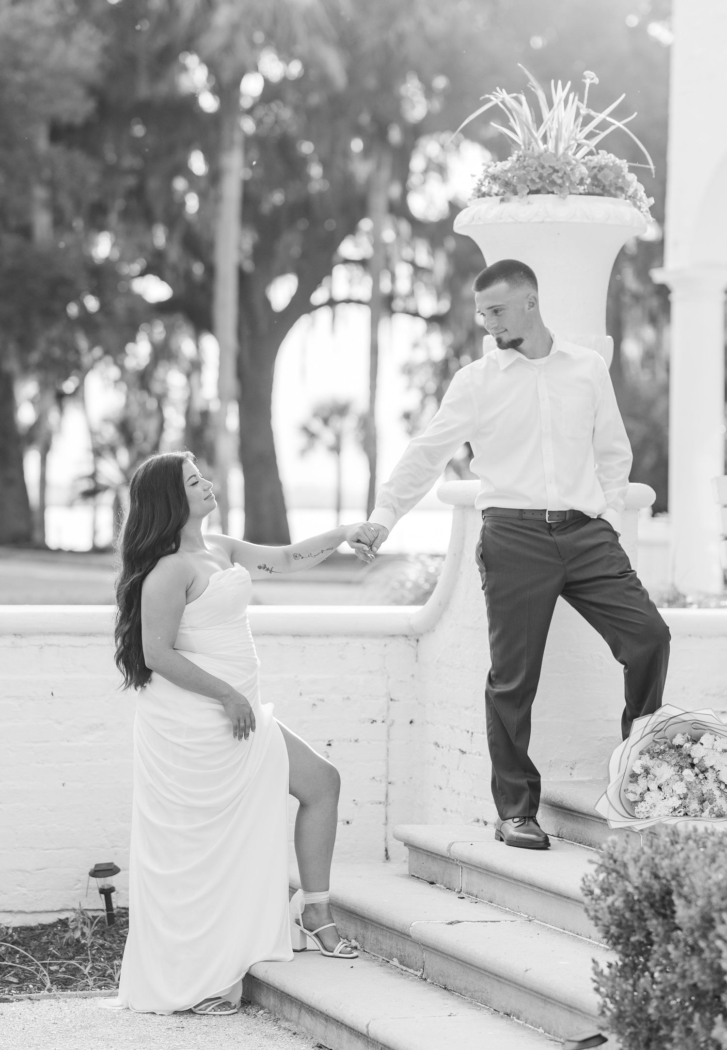 Black and white photo of a couple on outdoor steps, woman in white gown holds mans hand as he stands above her.