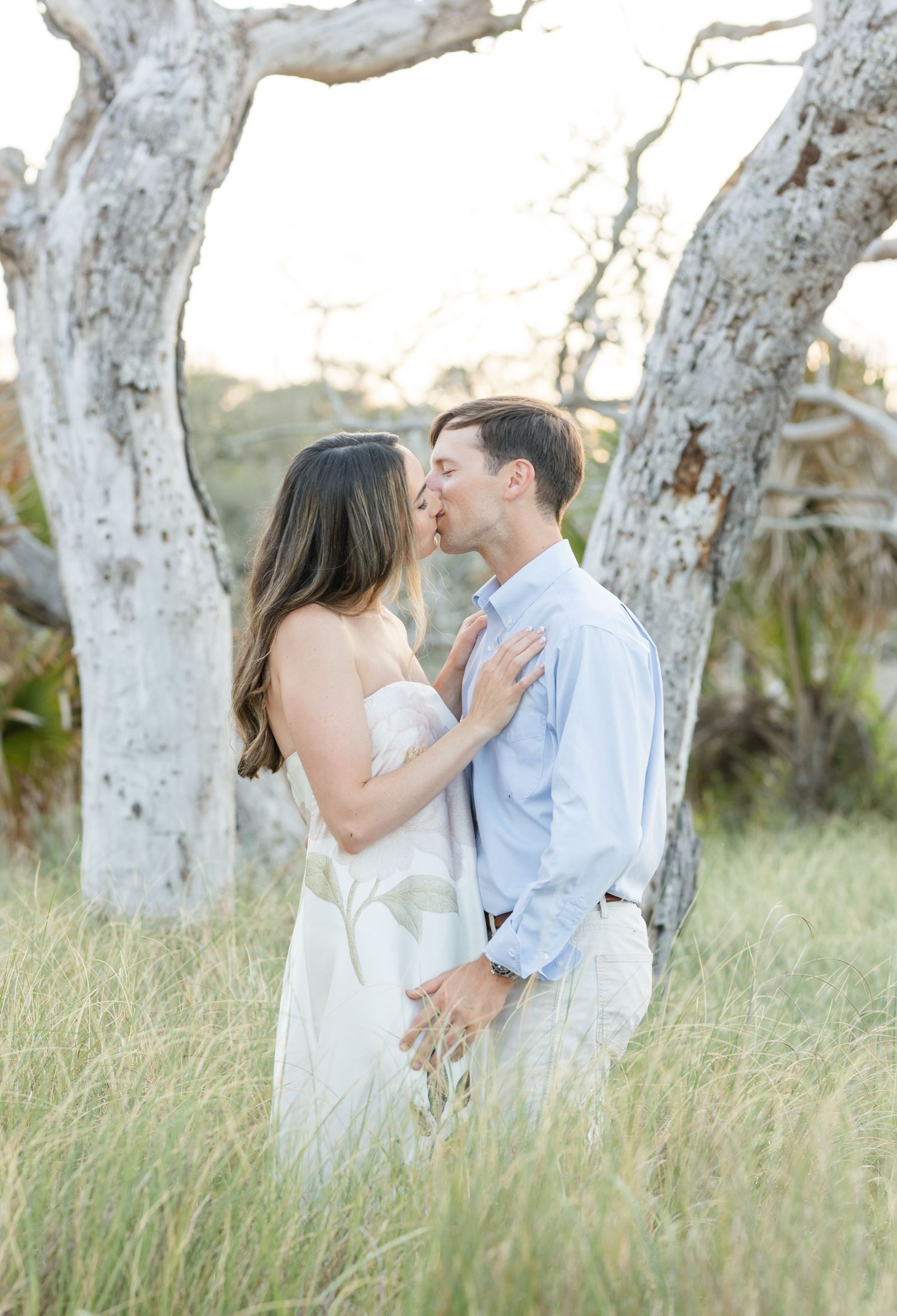 Couple sharing a romantic kiss in a golden field surrounded by white trees during an outdoor engagement session.