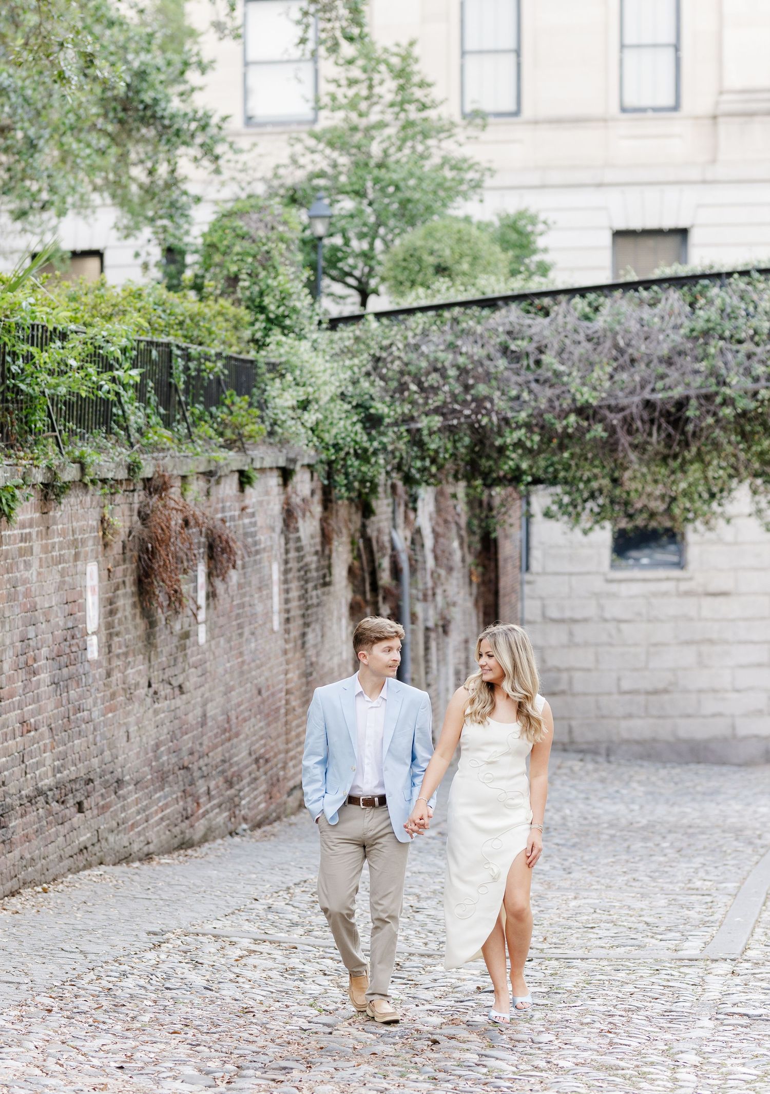Couple walks hand-in-hand on cobblestone street, man in blue blazer, woman in white dress, brick wall with greenery behind.