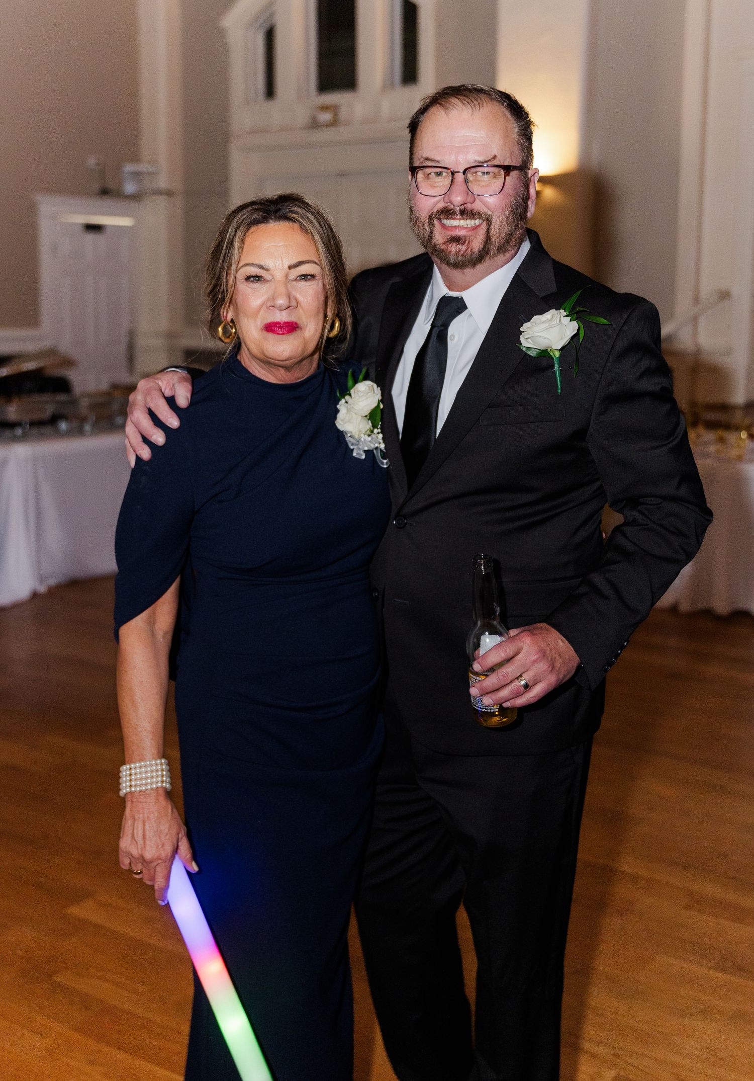 Couple in formal attire poses together at a wedding reception holding glow sticks.