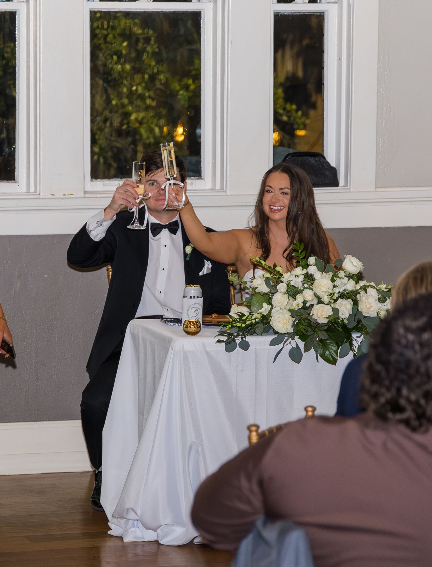 Bride and groom raising champagne glasses in a toast at their wedding reception beside a white floral centerpiece.