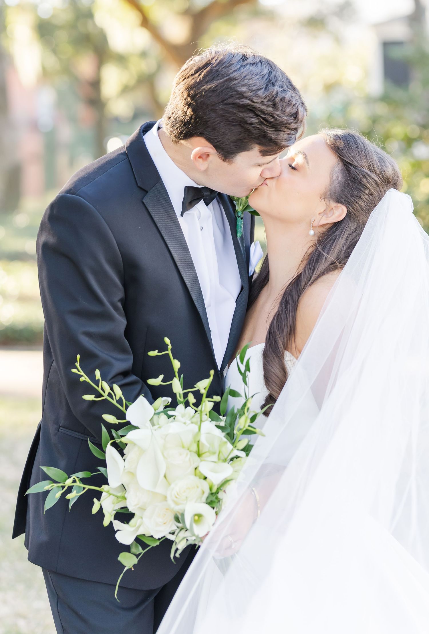 Bride and groom share a romantic kiss outdoors, bride holding white orchid bouquet with veil.