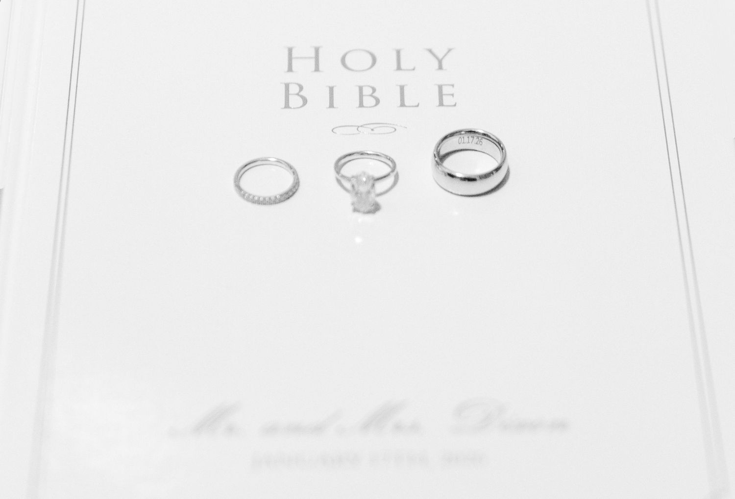 Three silver wedding rings displayed on a personalized Holy Bible, including a diamond engagement ring and two bands.