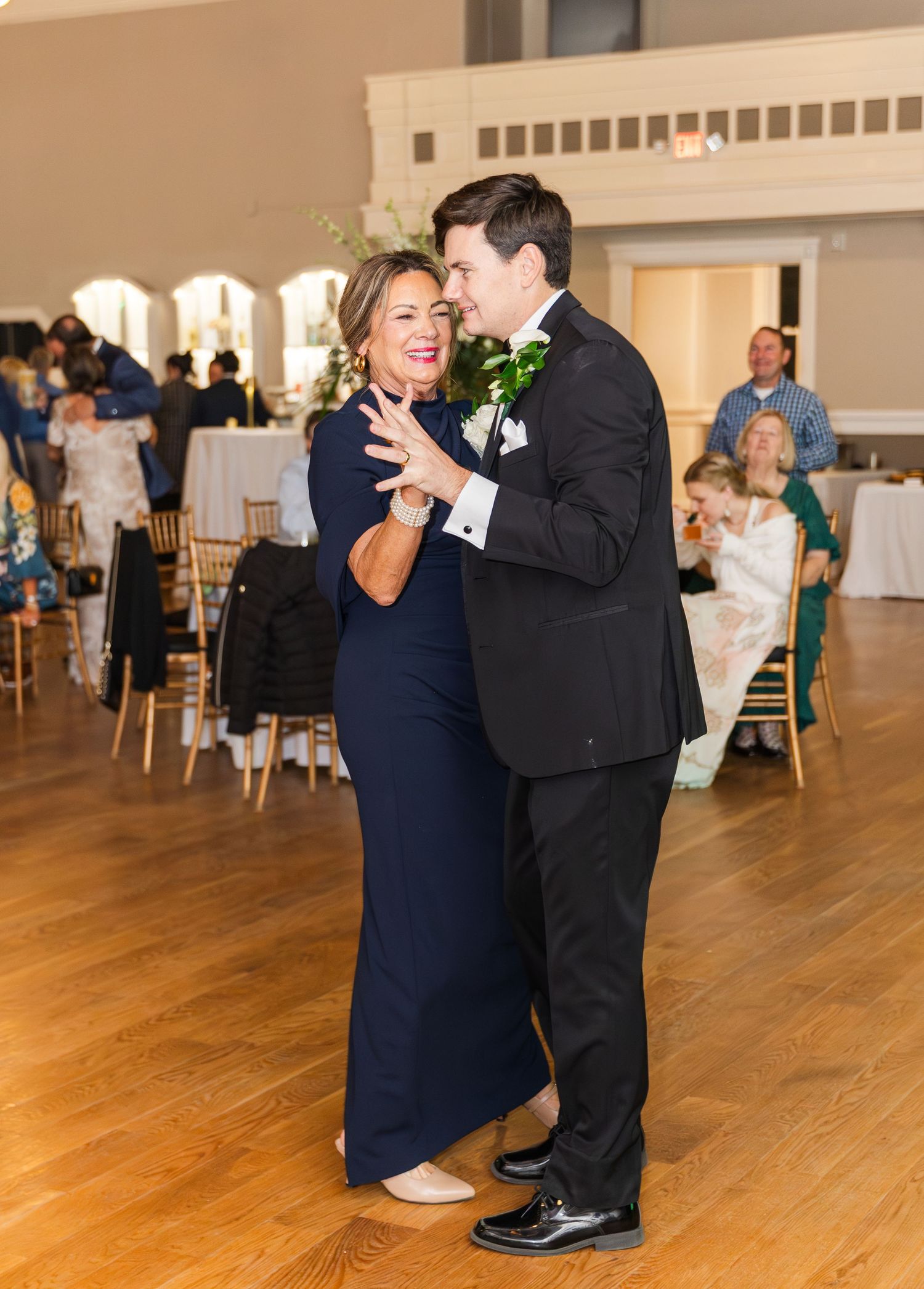 Groom in black tuxedo shares mother-son dance with woman in navy gown at wedding reception.