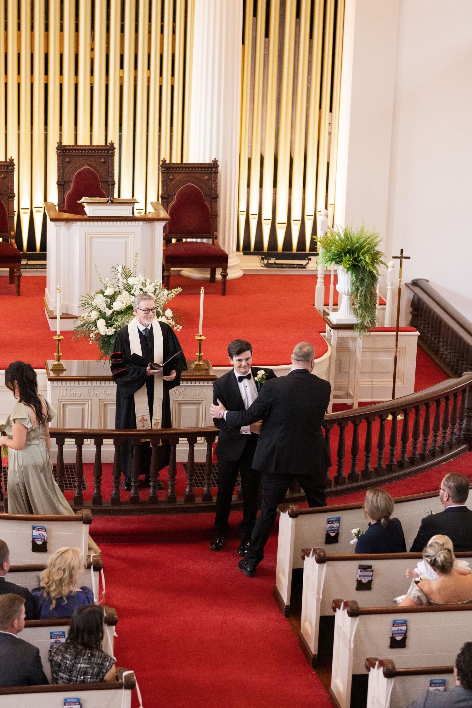 Groom greeting guests at church altar with officiant and bridesmaids during wedding ceremony.