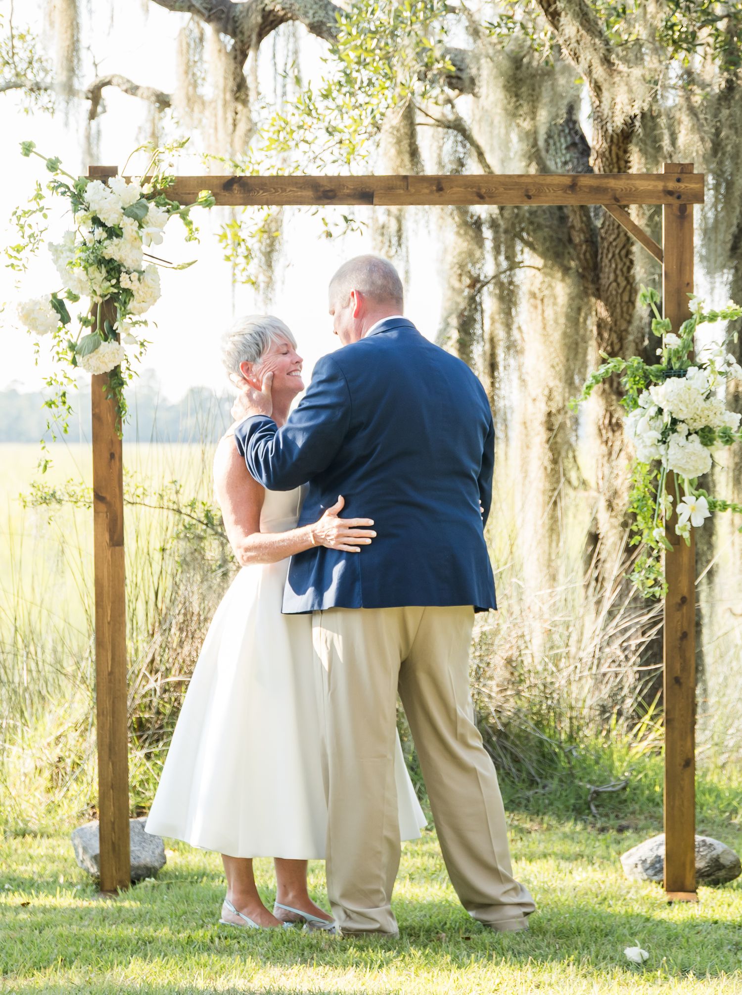 Couple embracing under a wooden arch decorated with white flowers and Spanish moss in background.